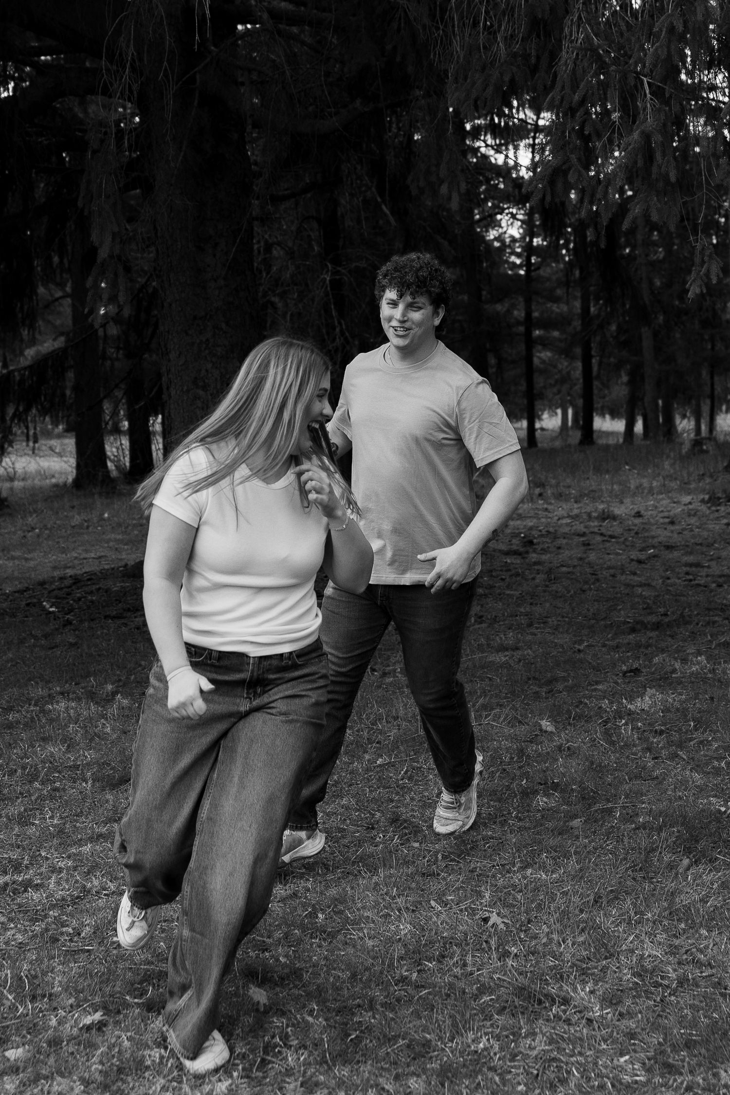 Black and white photo of a young couple walking and laughing together in a wooded park setting.