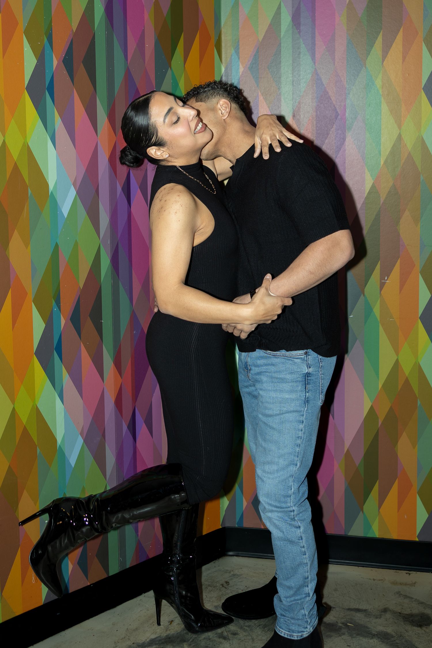 A couple embraces and kisses in front of a colorful geometric wall, woman in black dress with boots, man in jeans.