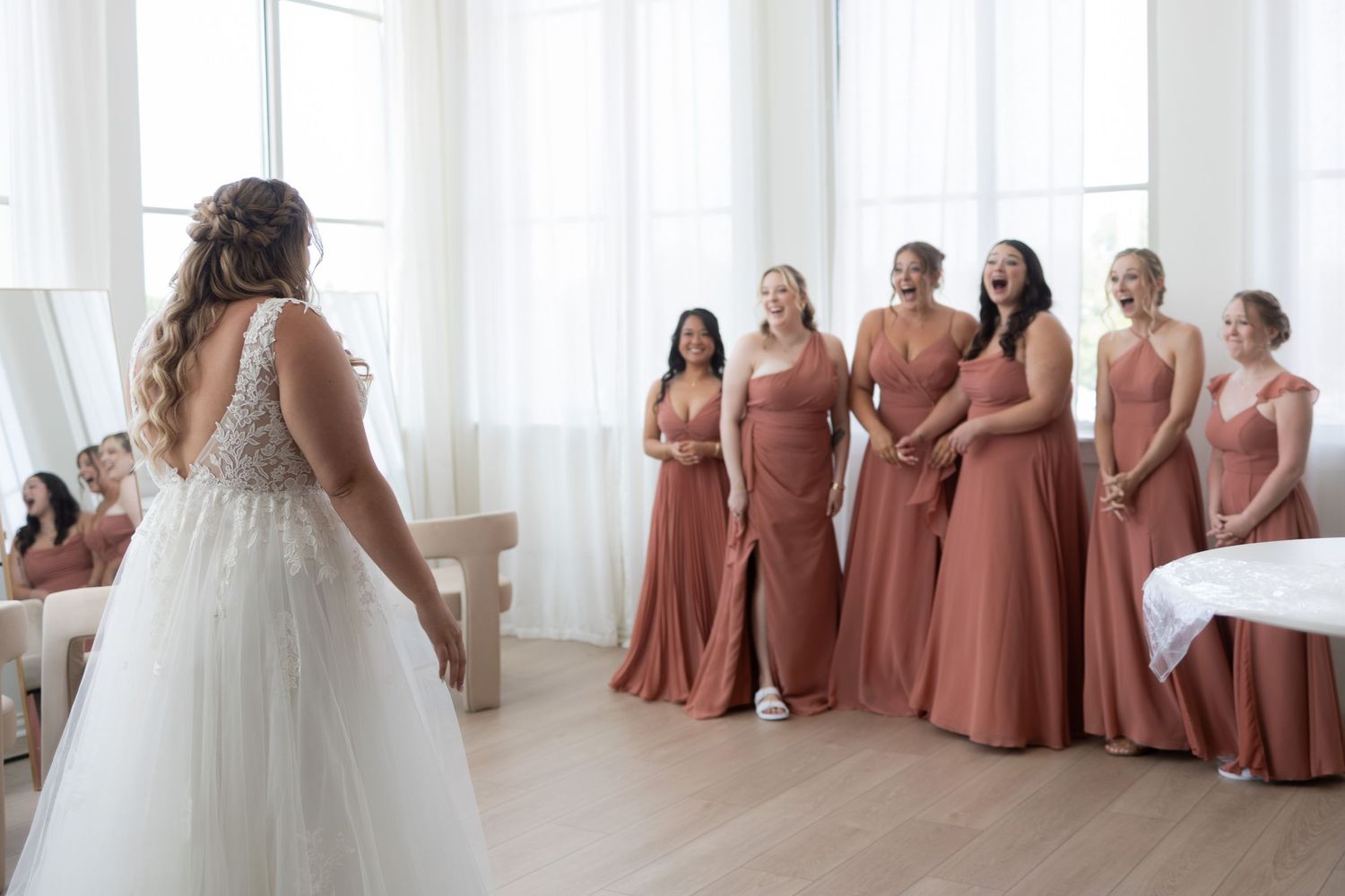 Bride in white lace gown shares first look moment with bridesmaids in dusty rose dresses in bright white room.