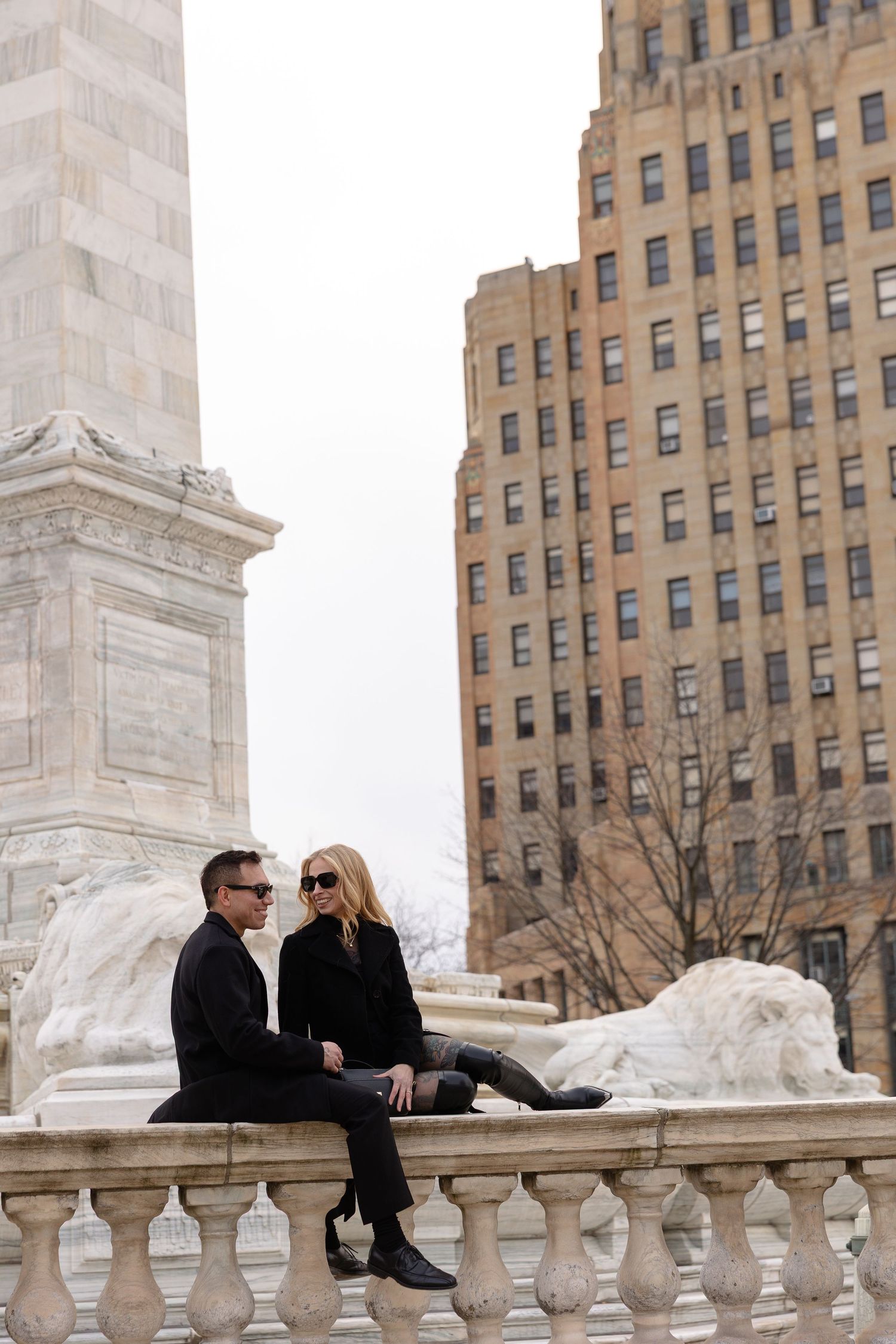 Couple in black outfits sitting on marble balustrade near lion sculpture and tall building near Buffalo City Hall
