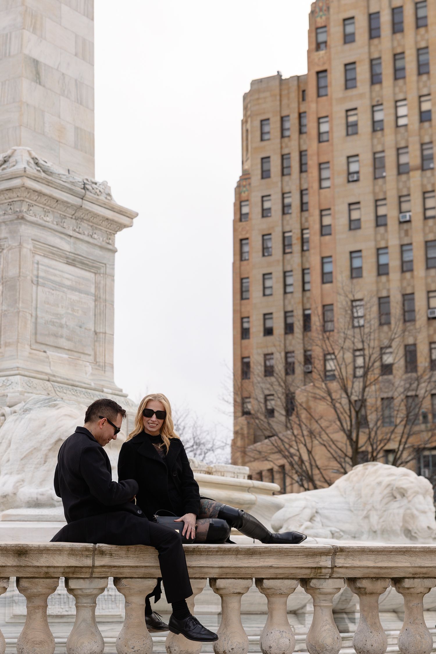 Two people in black outfits sitting on a marble balustrade near a monument by Buffalo City Hall