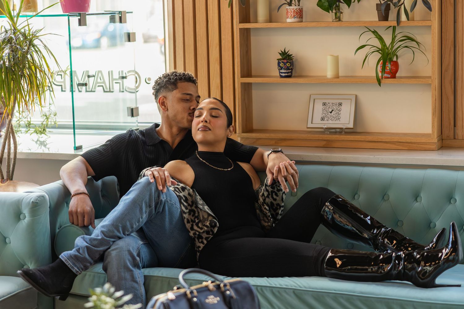 Couple relaxing on a teal sofa, man in black shirt and jeans, woman in leopard print top and black boots.