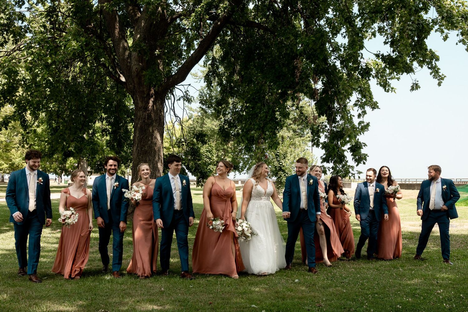 Wedding party in navy suits and rust bridesmaid dresses posing under a large tree on green lawn.