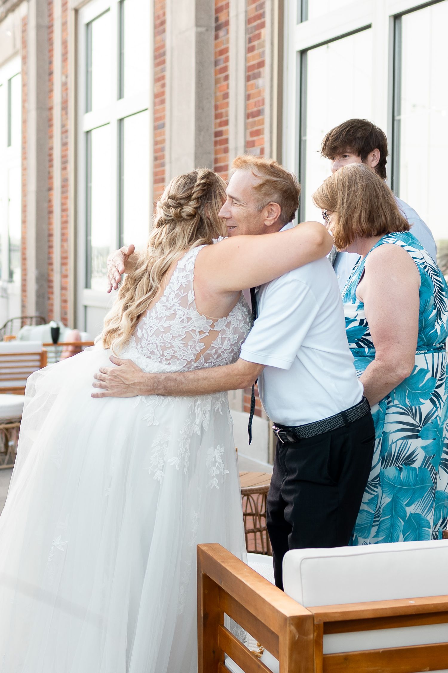 Bride in lace gown embracing a smiling man at an outdoor wedding reception near a brick building.
