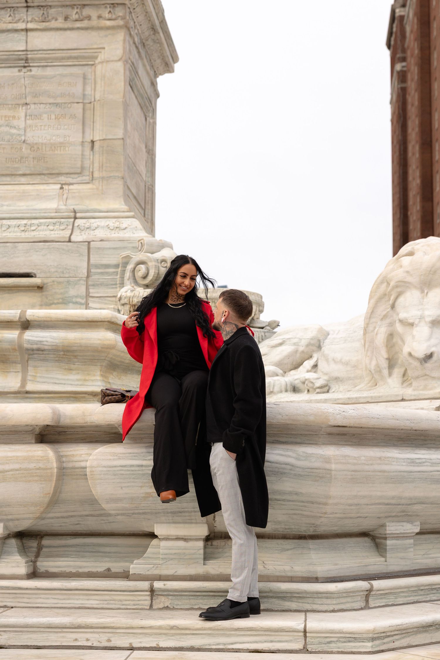 A couple in elegant black outfits, woman in red coat, posing by marble lion sculpture at a grand monument.