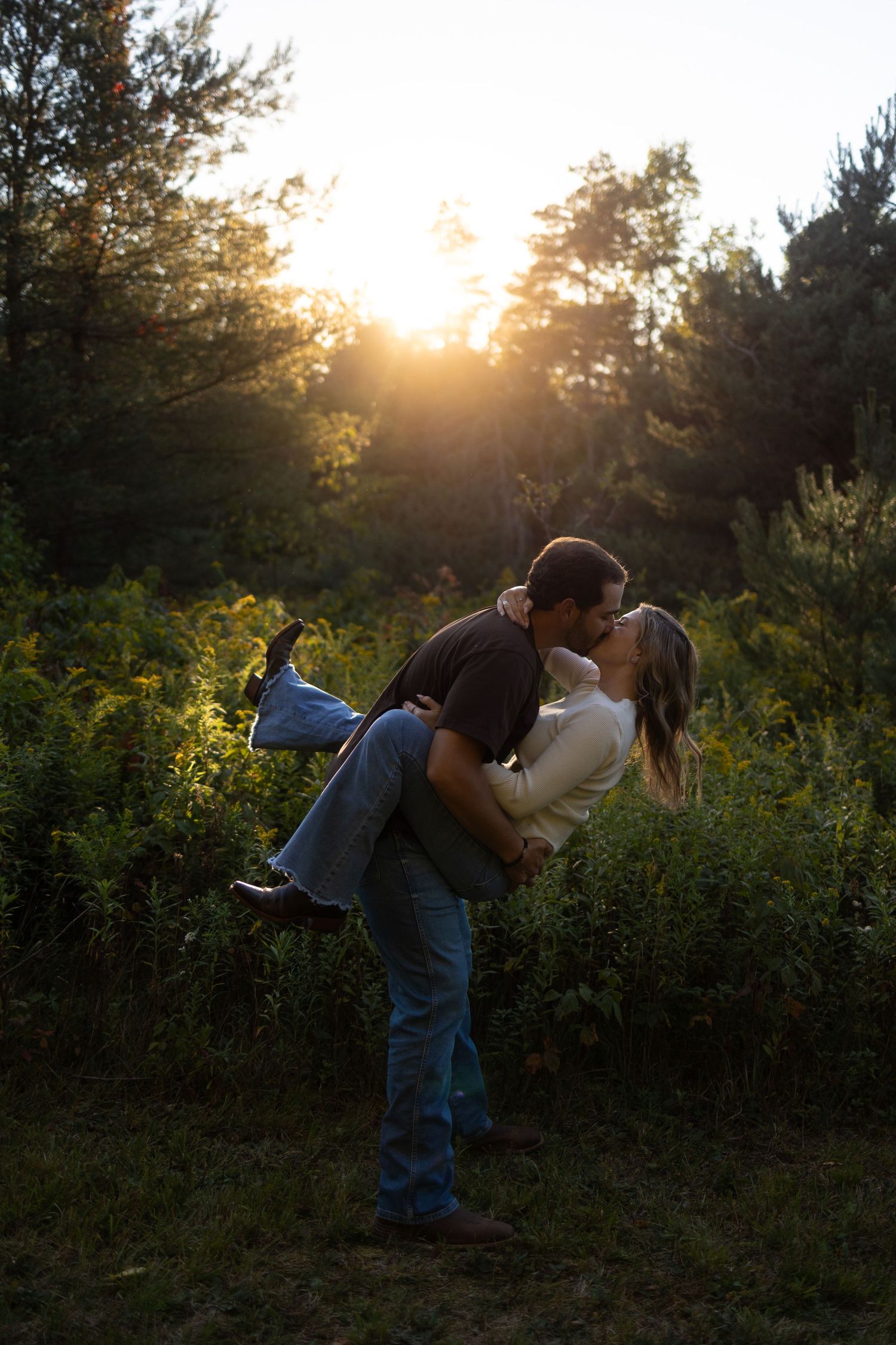 Couple sharing a romantic kiss at sunset in a lush green forest, man lifting woman off the ground.