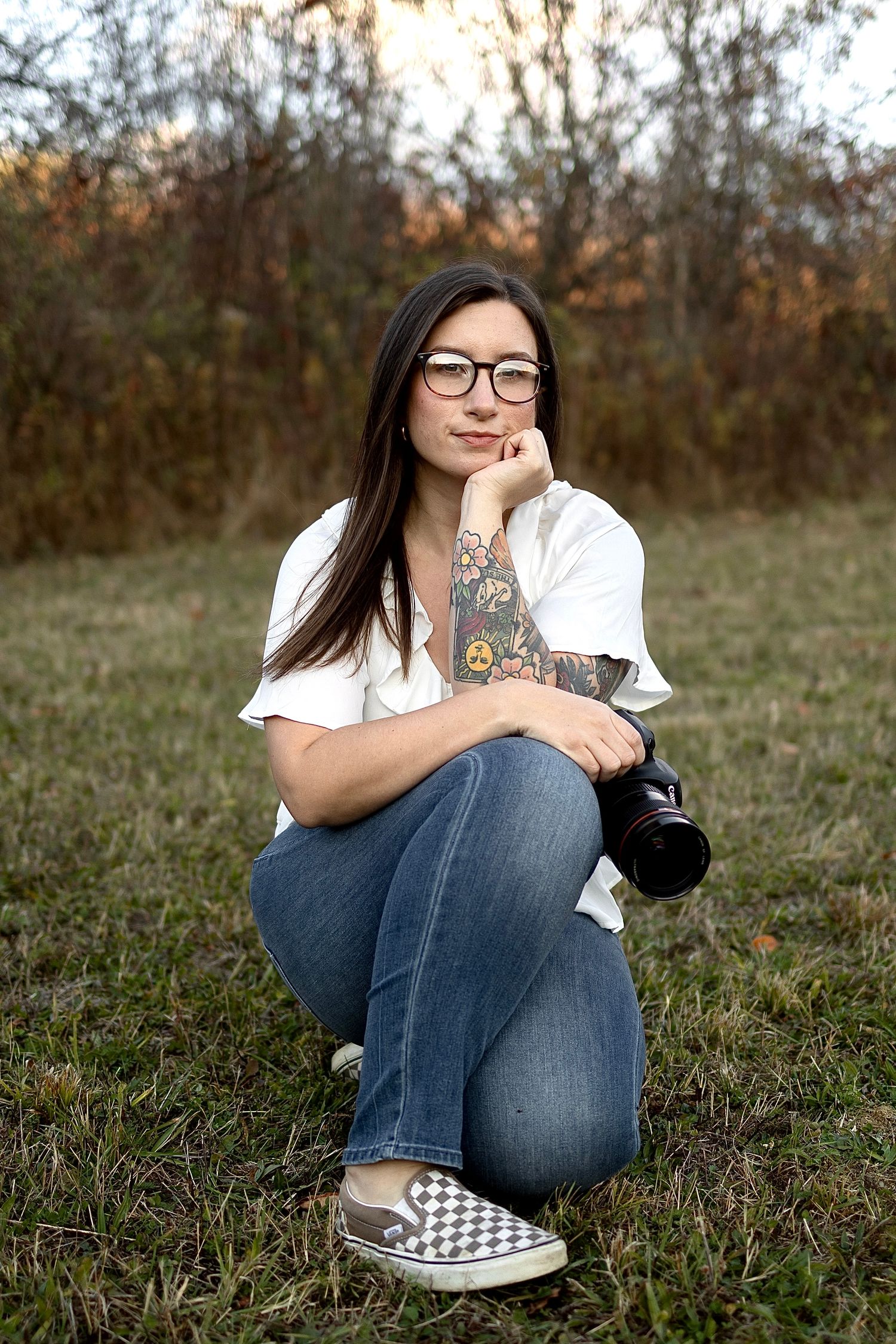 Young woman with tattoos and glasses crouching outdoors holding a camera, wearing jeans and a white shirt.