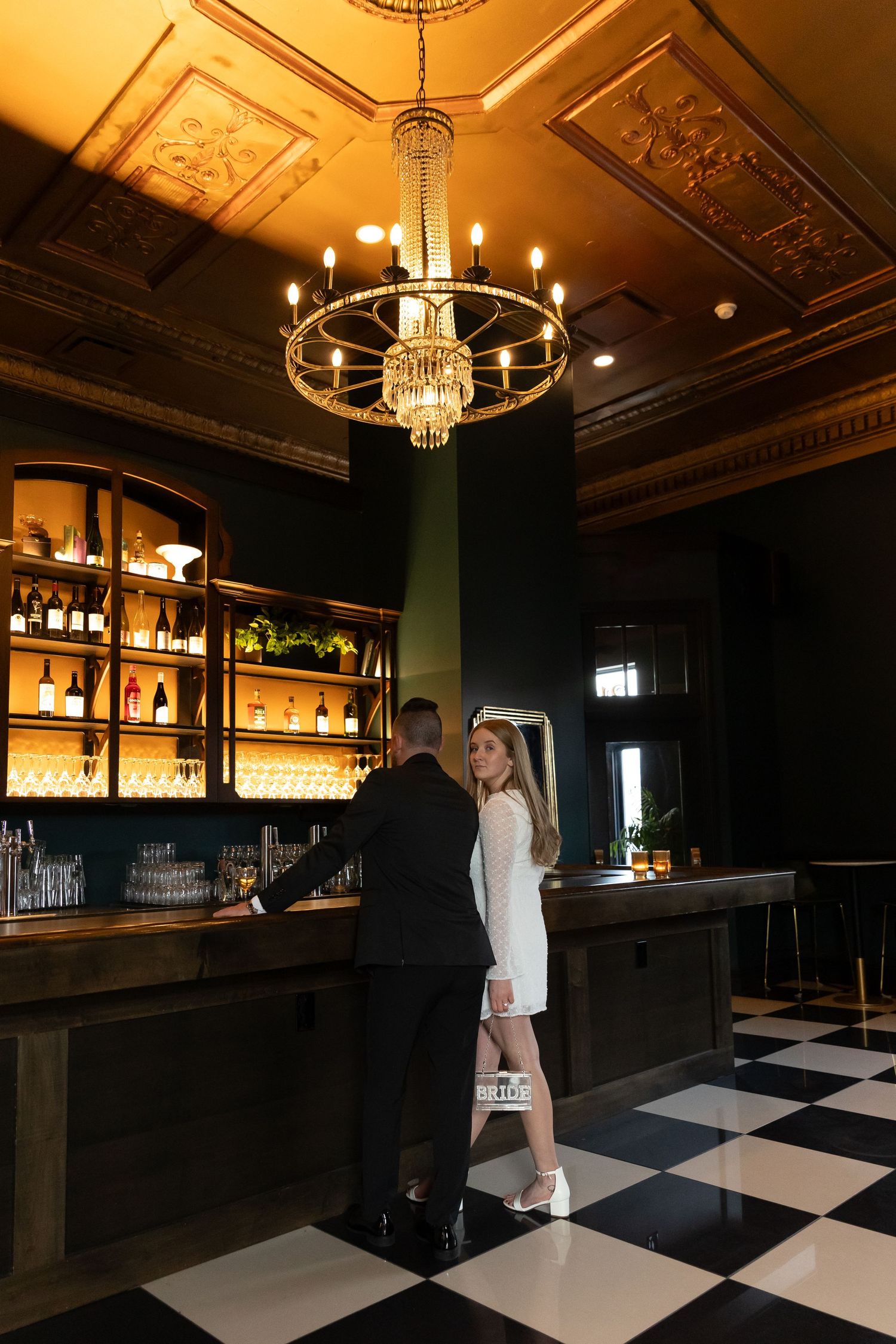 Couple in elegant dark bar with chandelier, black and white checkered floor, and backlit shelves of bottles.