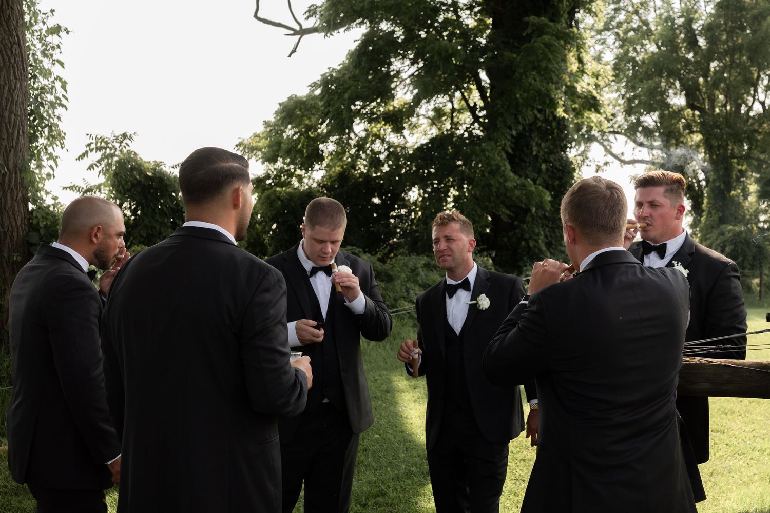 Groomsmen in black tuxedos with white boutonnieres gather outdoors under trees, adjusting their attire after their wedding ceremony.