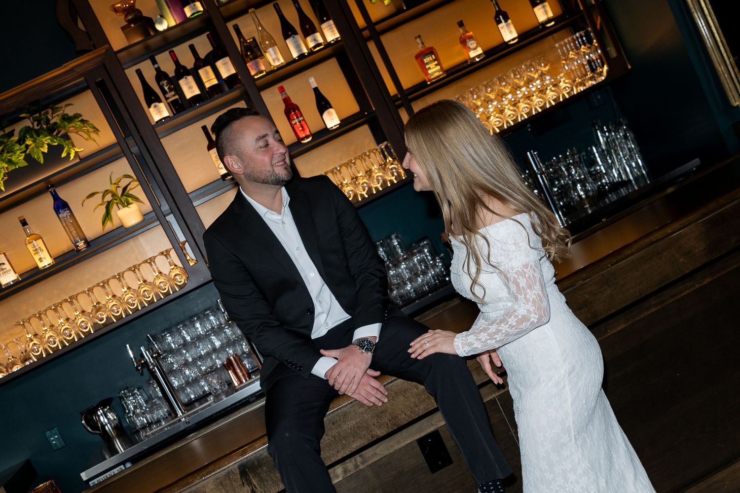 Couple sharing a romantic moment at an elegant bar with glowing shelves of bottles and glassware.