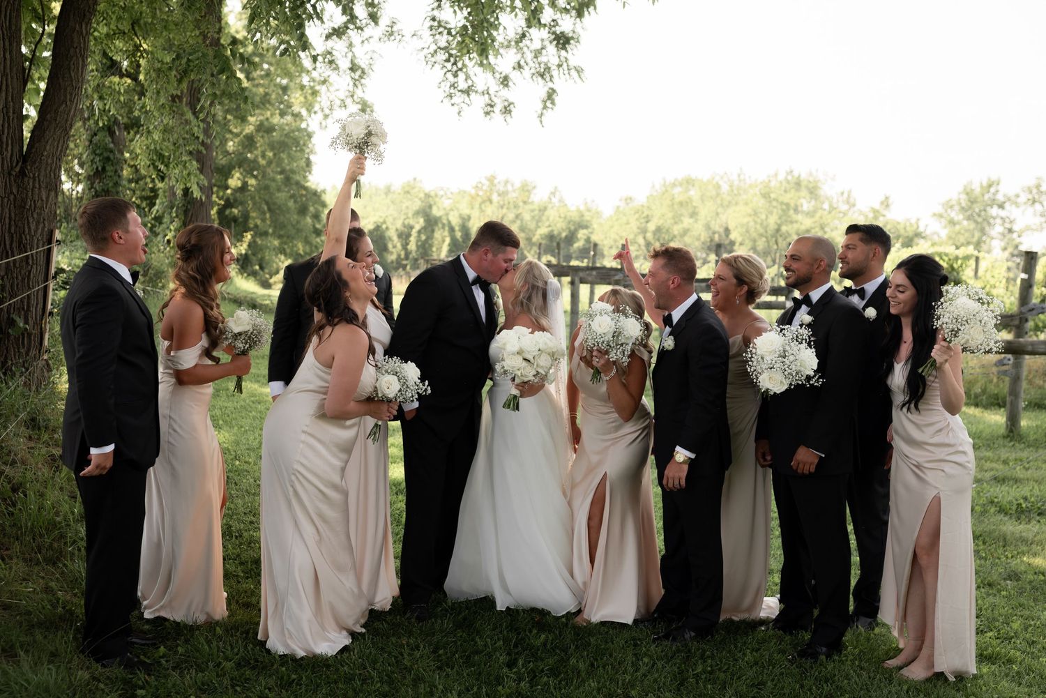 Wedding party celebrating outdoors, bride and groom kissing surrounded by bridesmaids in champagne gowns and groomsmen in black suits.