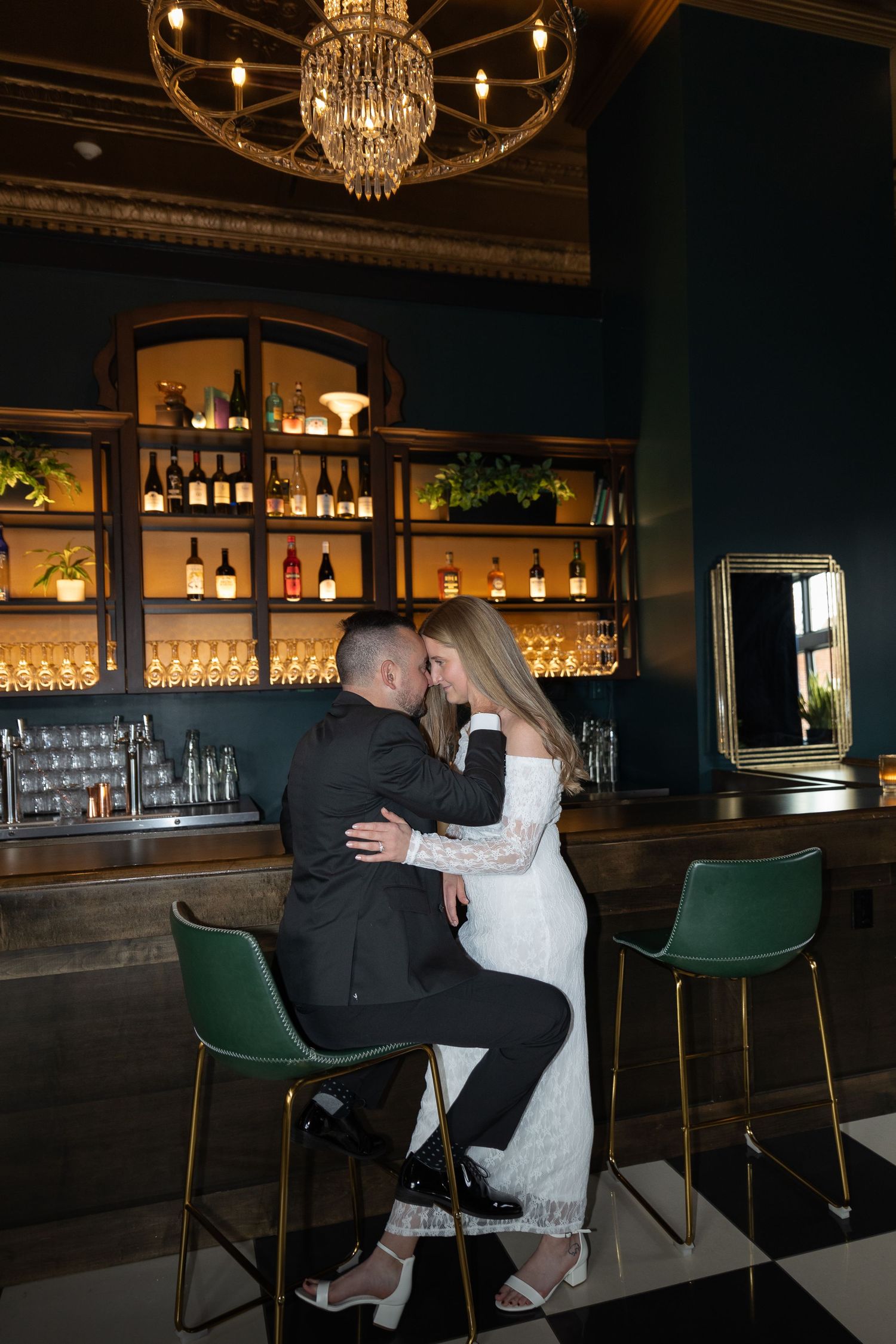 Couple sharing a romantic moment at an elegant bar with green velvet stools, bottle-lined shelves, and a chandelier above.
