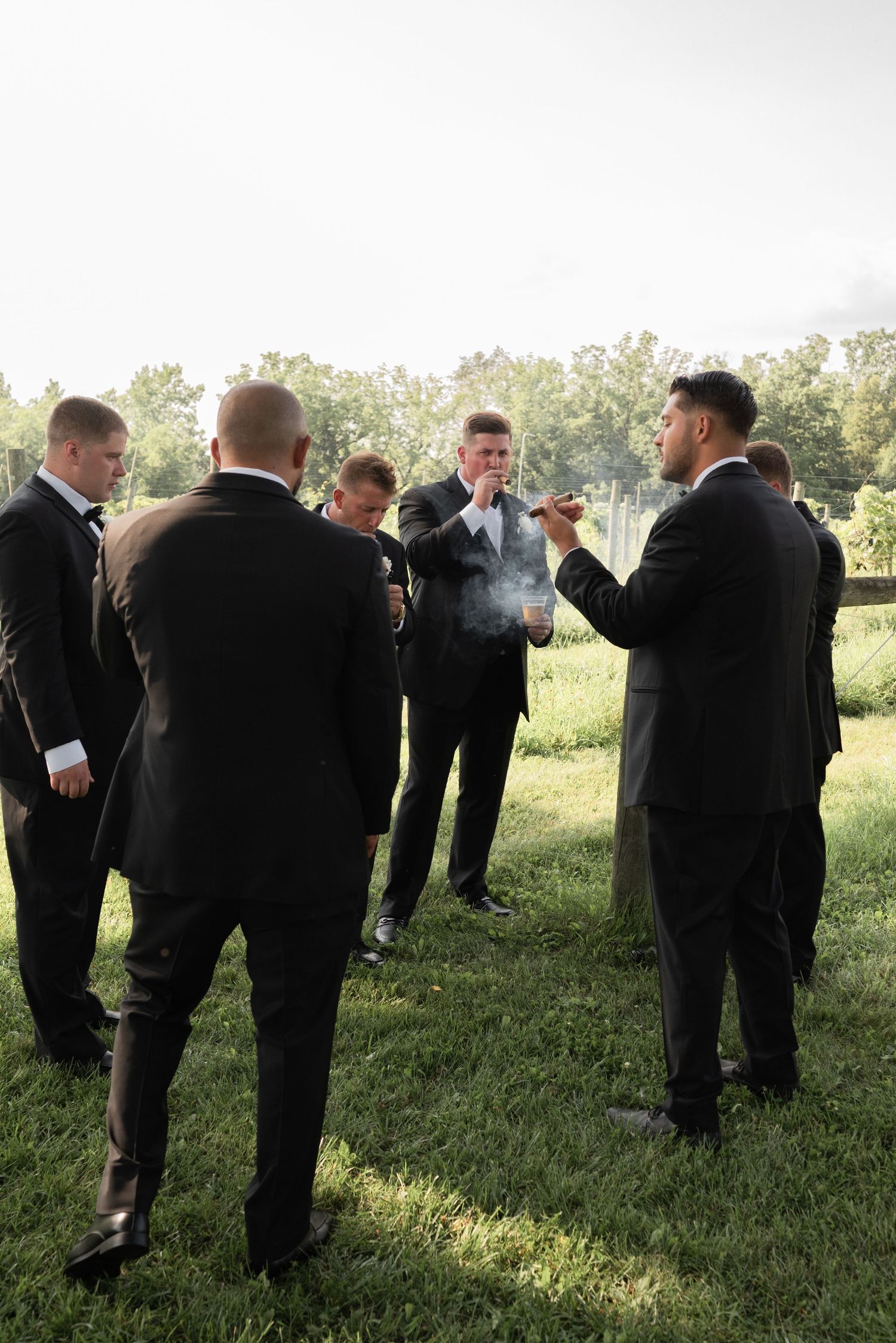 Groomsmen in black suits smoking cigars outdoors on green grass before a wedding ceremony in Rochester, NY