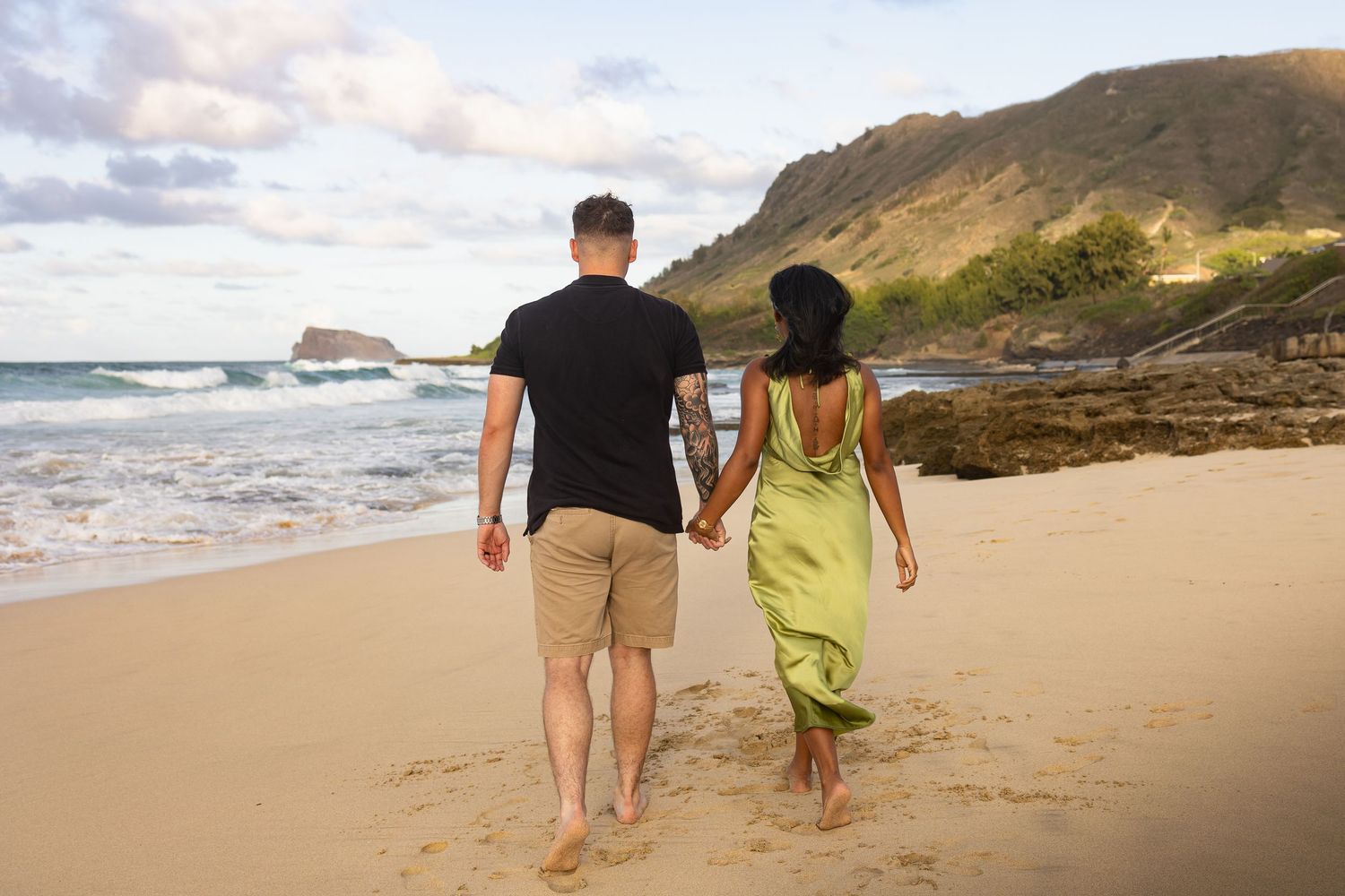 Couple holding hands walking barefoot on a sandy beach in Hawaii at sunset with ocean waves and green mountains in background