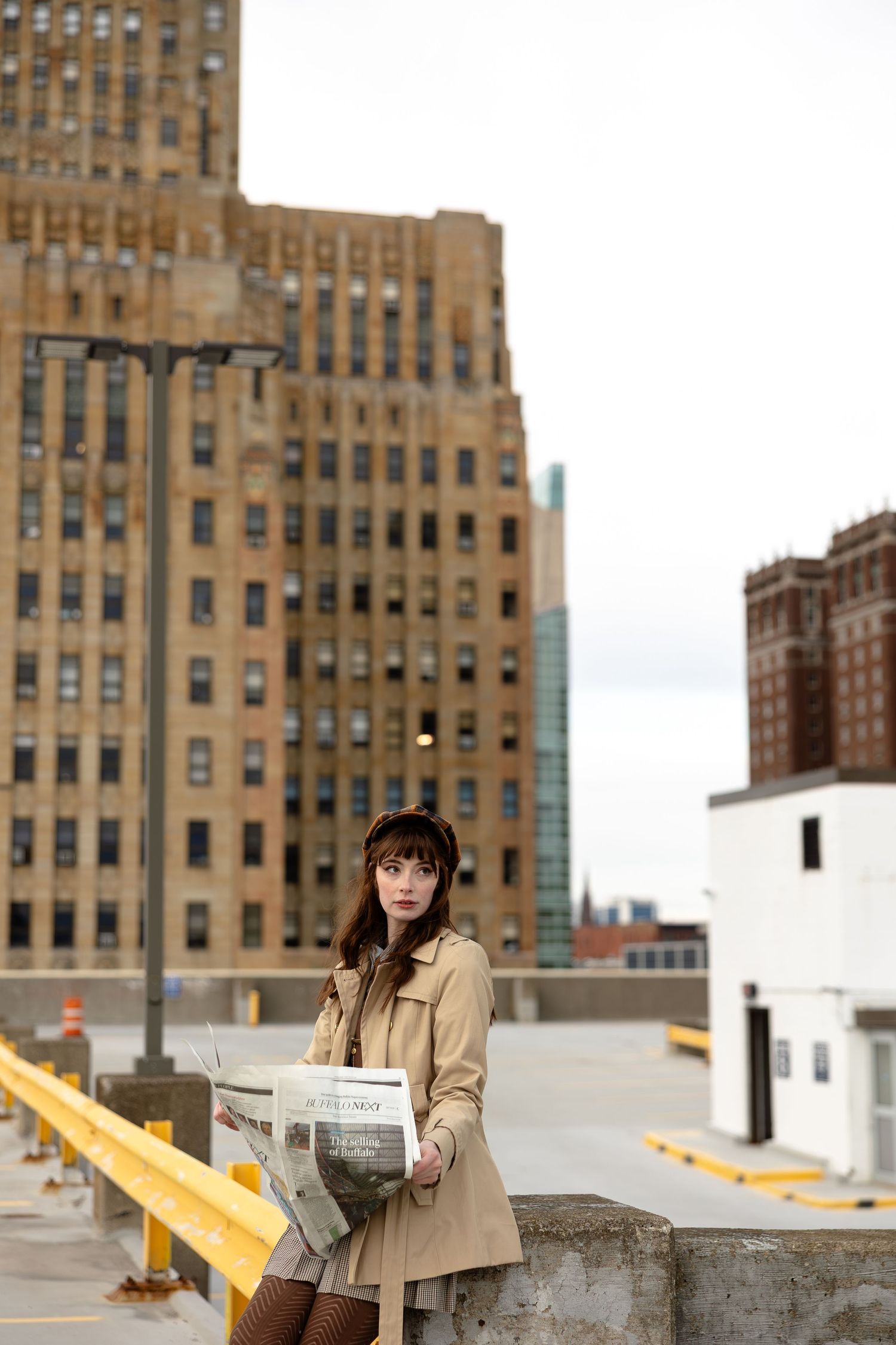 Young woman in trench coat holding newspaper on urban rooftop parking garage with city buildings backdrop.