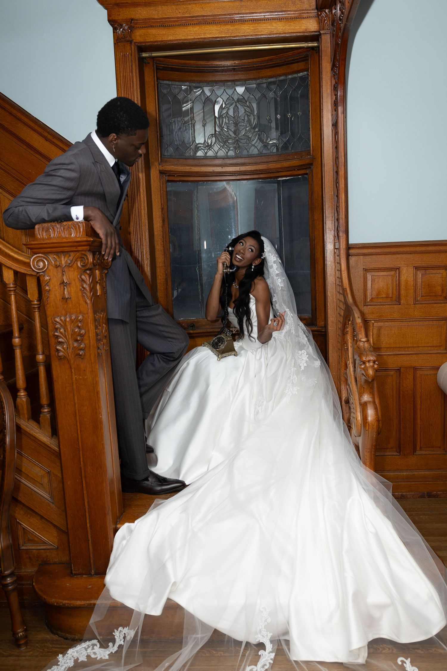 Black bride in white ball gown and veil laughs with groom in gray suit on ornate wooden staircase.