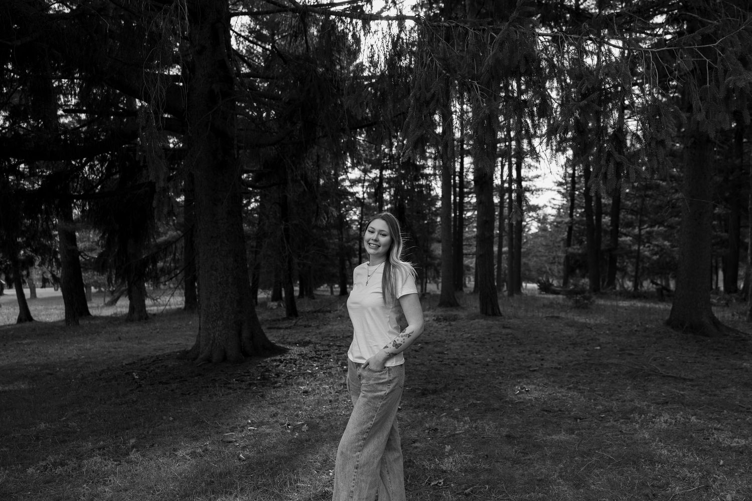 Young woman smiling in a black and white forest photo, standing among tall pine trees on a grassy woodland path.