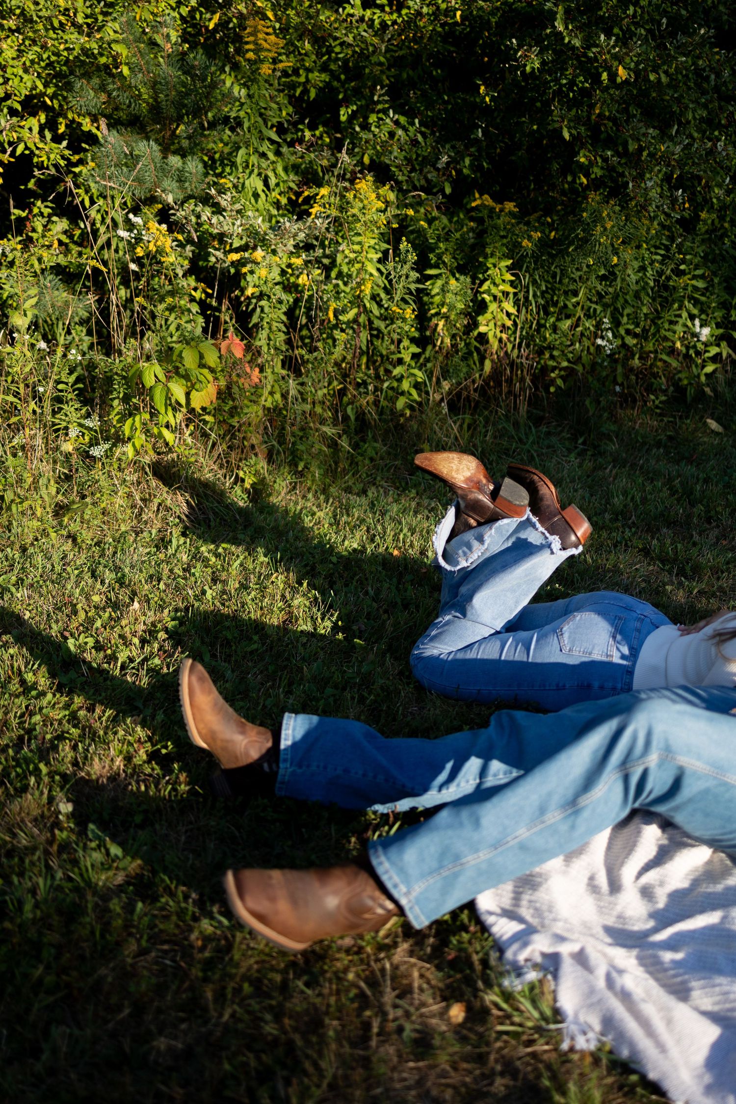 Two people wearing cowboy boots and blue jeans relaxing on green grass near lush bushes outdoors.