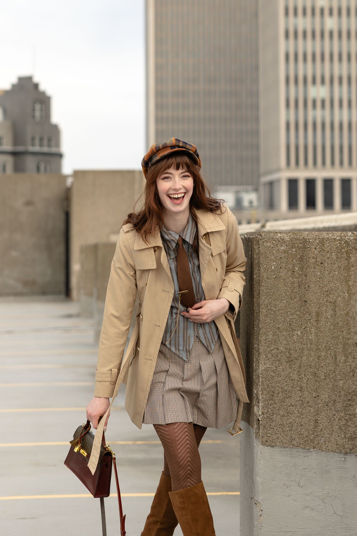 Young woman in beige trench coat, plaid cap, brown boots, and fishnet tights poses on rooftop with city skyline backdrop.