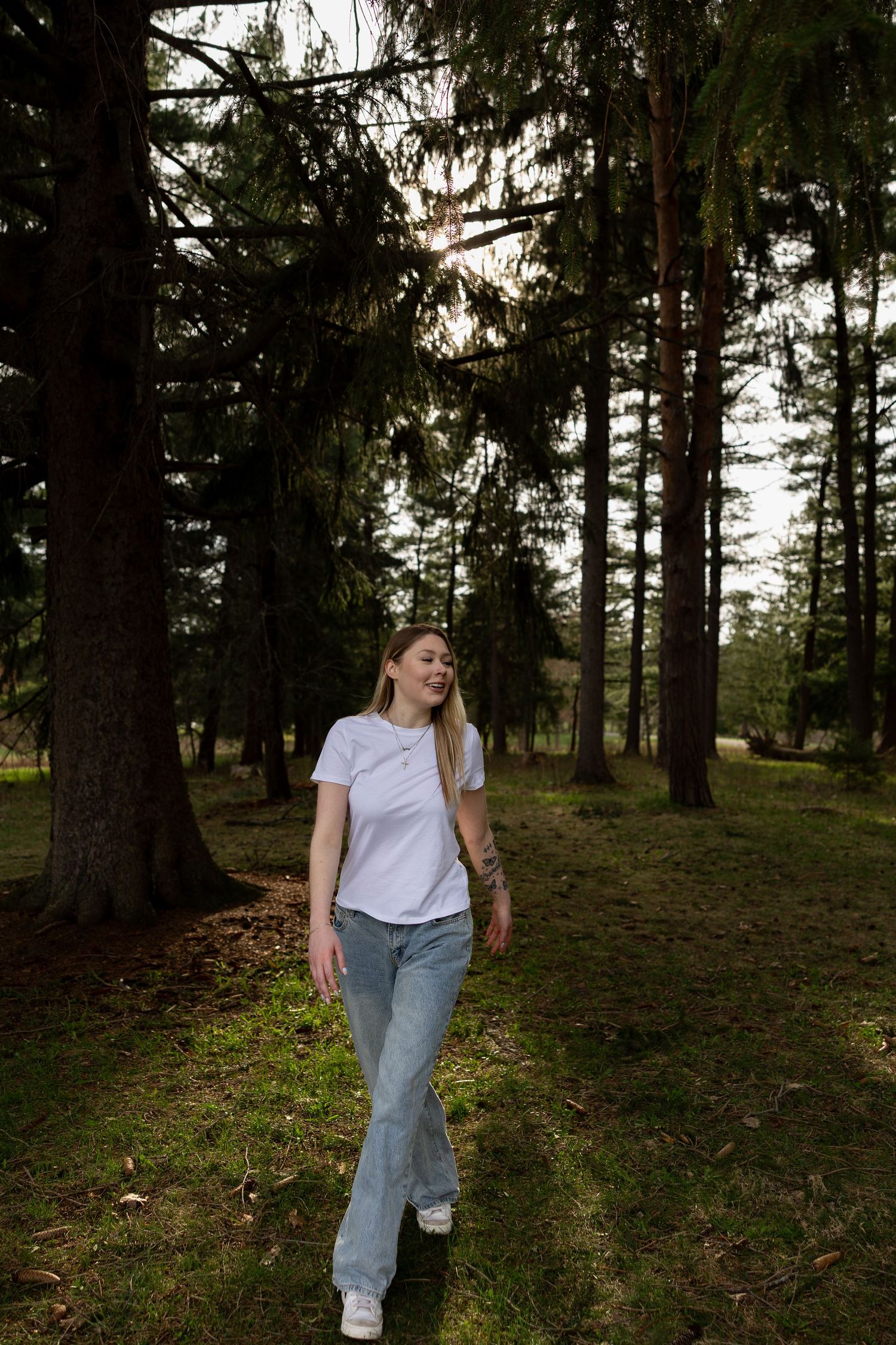 Young woman in white t-shirt and jeans walking through a sunlit forest with tall pine trees.