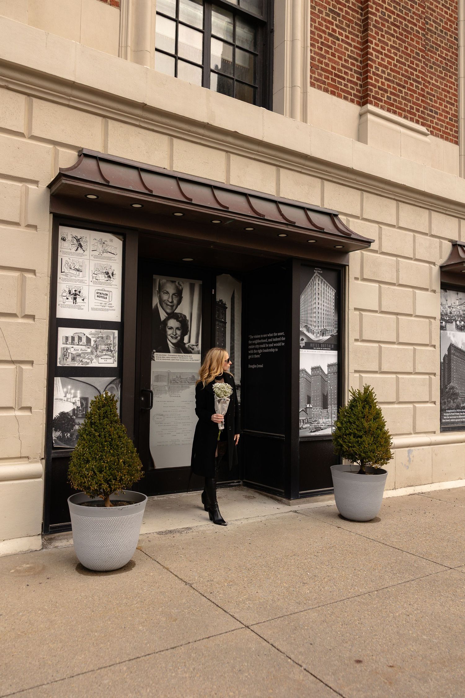 Woman in black outfit viewing historical photo display outside brick building with potted topiary plants.