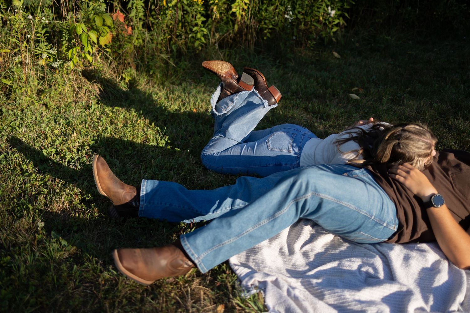 Two people lying on green grass outdoors, wearing jeans and brown boots, relaxing in natural sunlight.