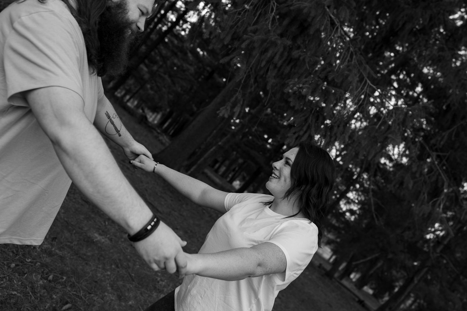 Black and white photo of a couple holding hands outdoors, woman leaning back playfully among trees.