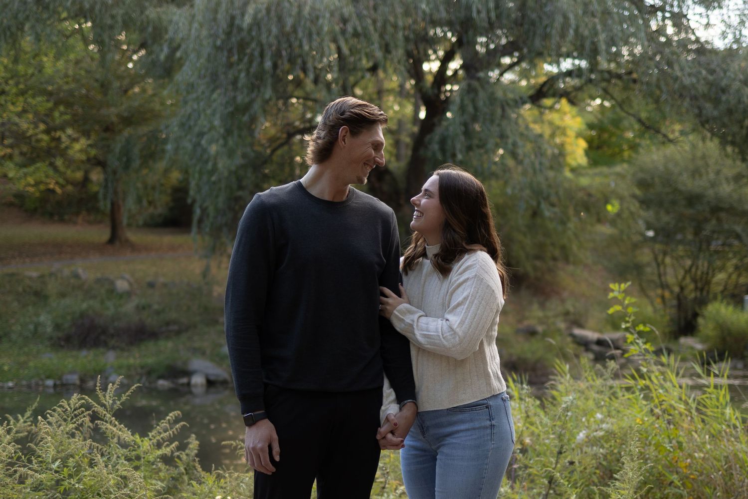 A young couple holds hands and gazes at each other in a lush green park setting on a warm autumn day.