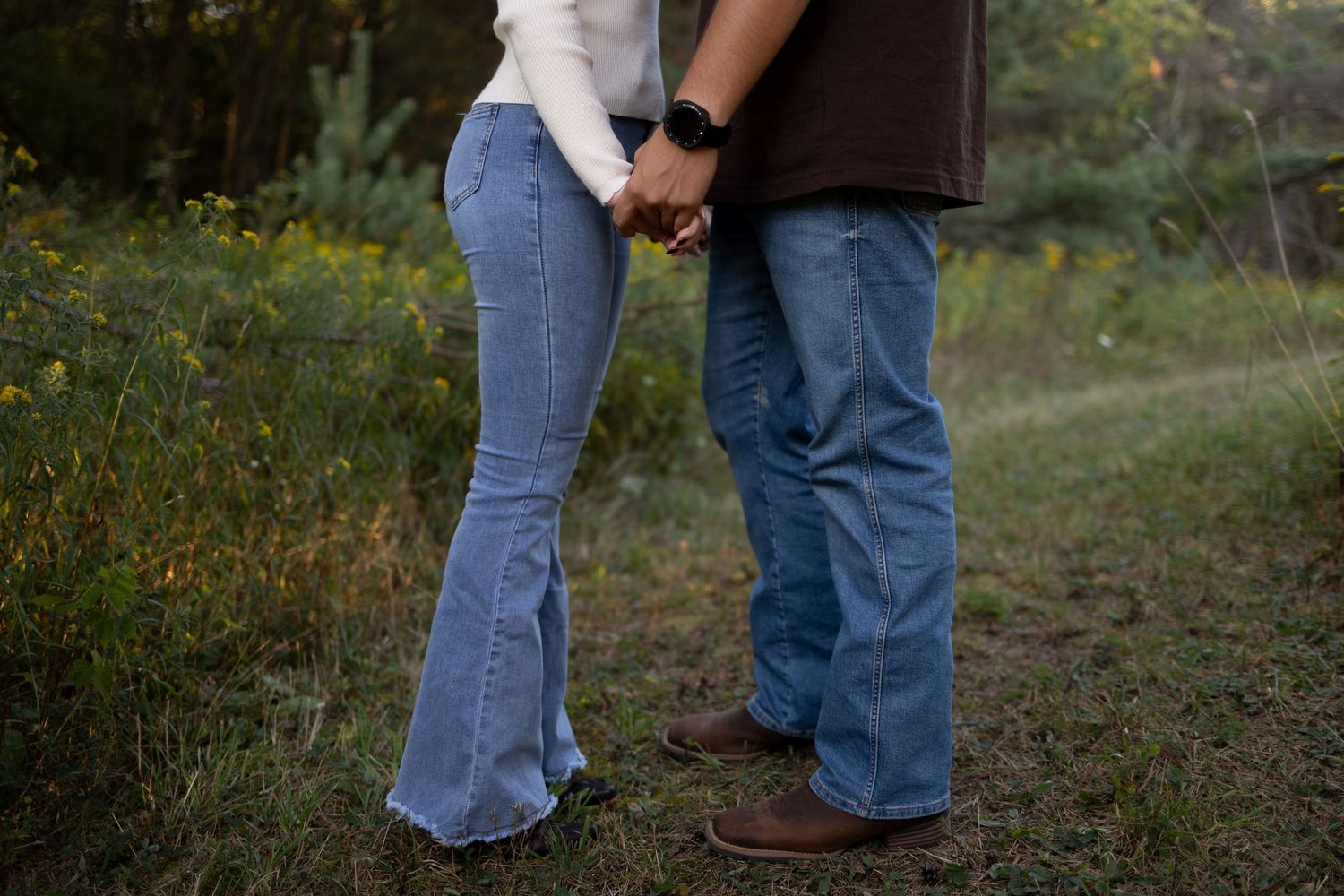 Couple holding hands in jeans and boots standing in a grassy field with wildflowers in background.