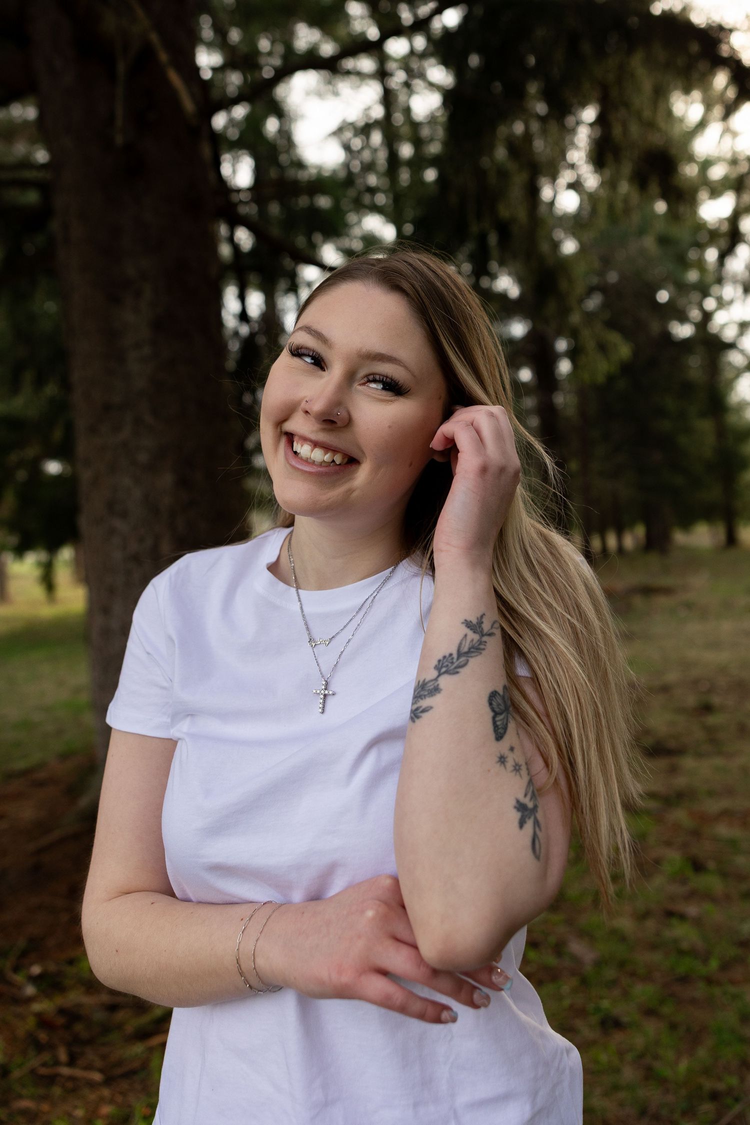 Smiling young woman with blonde hair and floral arm tattoo wearing white t-shirt and cross necklace in a park.