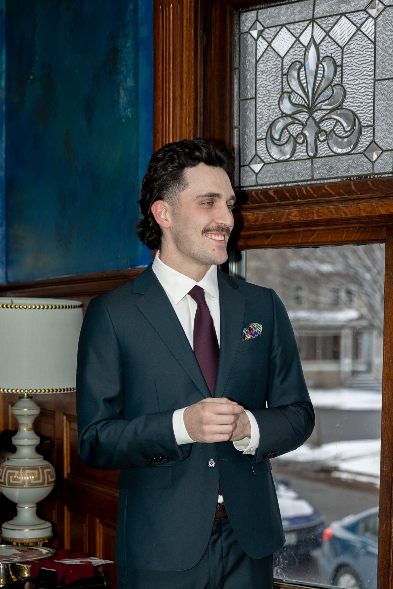 A well-dressed man in a dark suit and burgundy tie stands smiling near a stained glass window in an elegant interior.