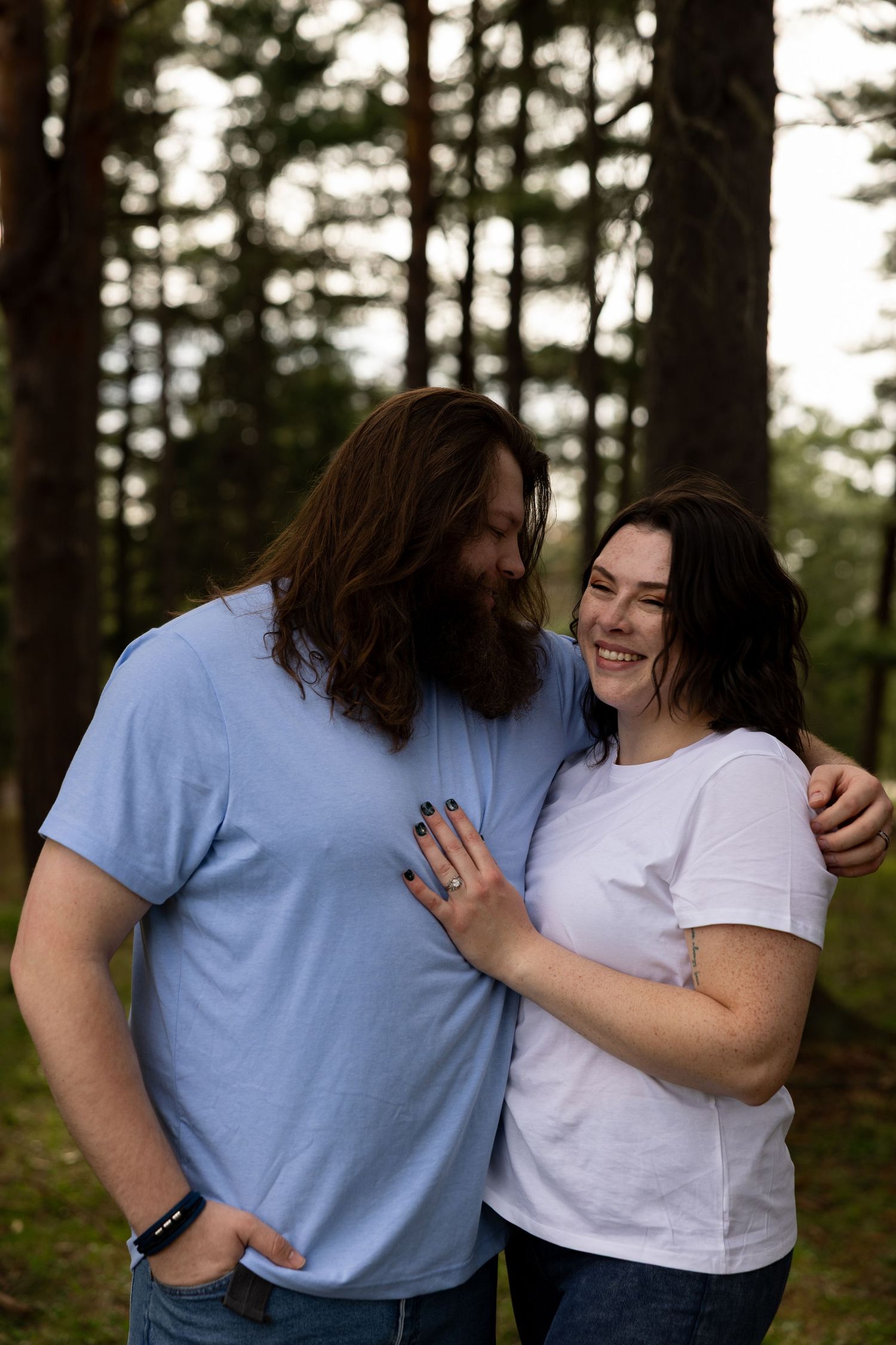 A couple smiles and embraces in a forest, man in blue tee with beard, woman in white tee showing engagement ring.