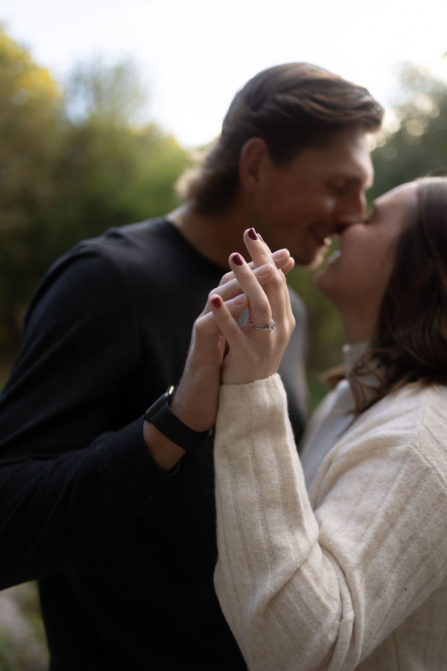 Couple sharing a romantic kiss outdoors, holding hands with an engagement ring visible, wearing black and cream sweaters.