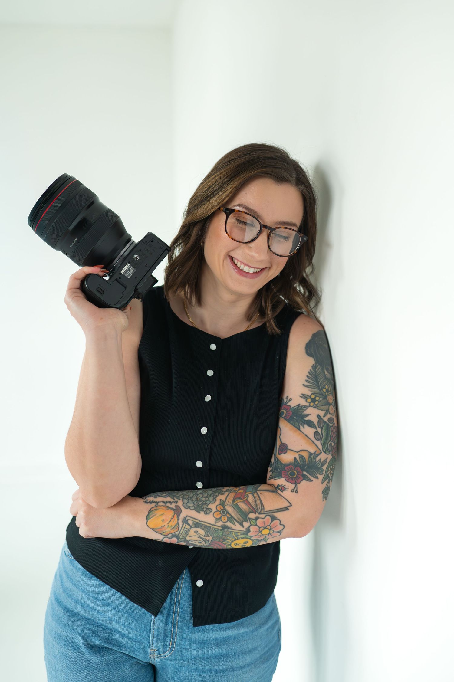 Smiling wedding and couples photographer with tattoos and glasses holding a mirrorless camera, wearing a black top and jeans.