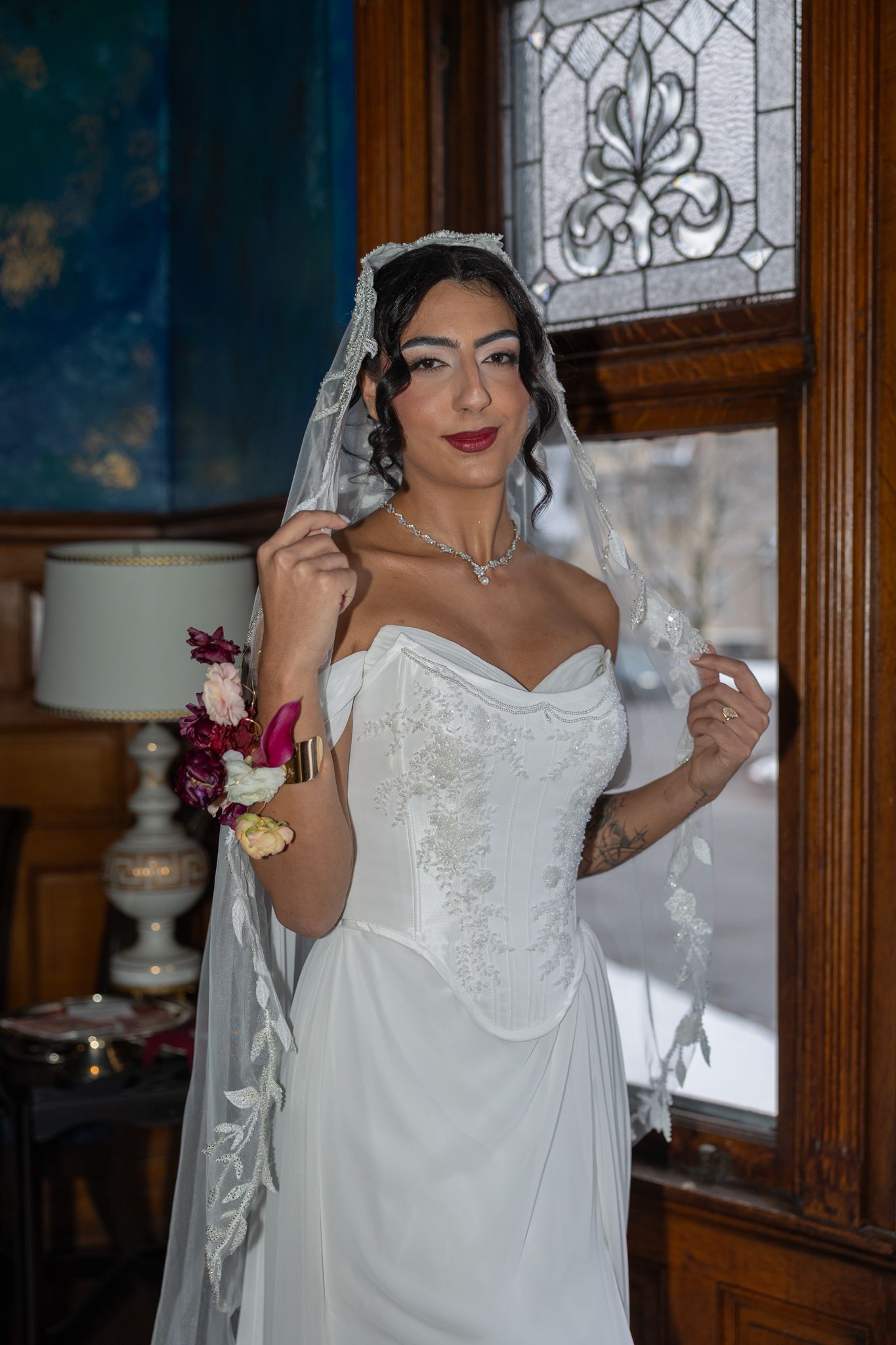 Bride in white off-shoulder gown with lace veil and bouquet poses by stained glass window in elegant interior.