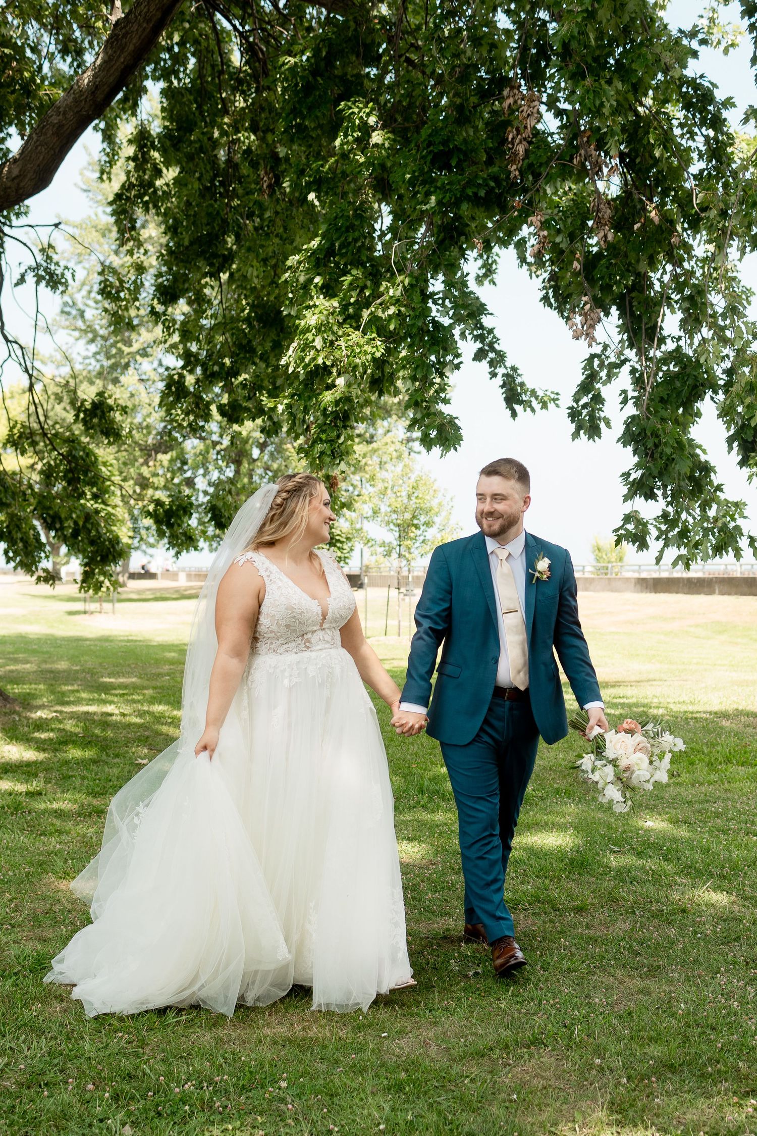 Bride and groom walk hand-in-hand under a lush green tree on their wedding day, smiling at each other outdoors.
