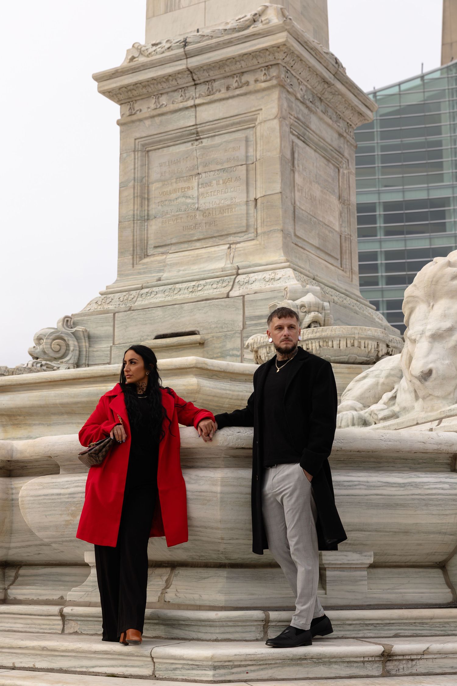 Couple posing at a marble monument, woman in red coat and man in black coat holding hands on stone steps.