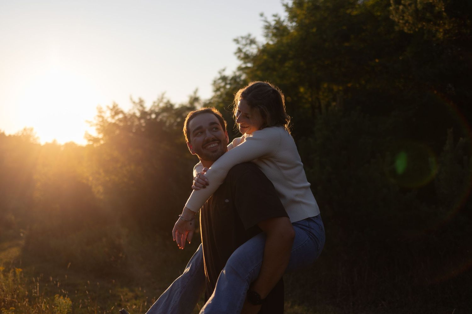 Couple sharing a romantic piggyback ride outdoors at golden hour sunset with lush green trees in background.