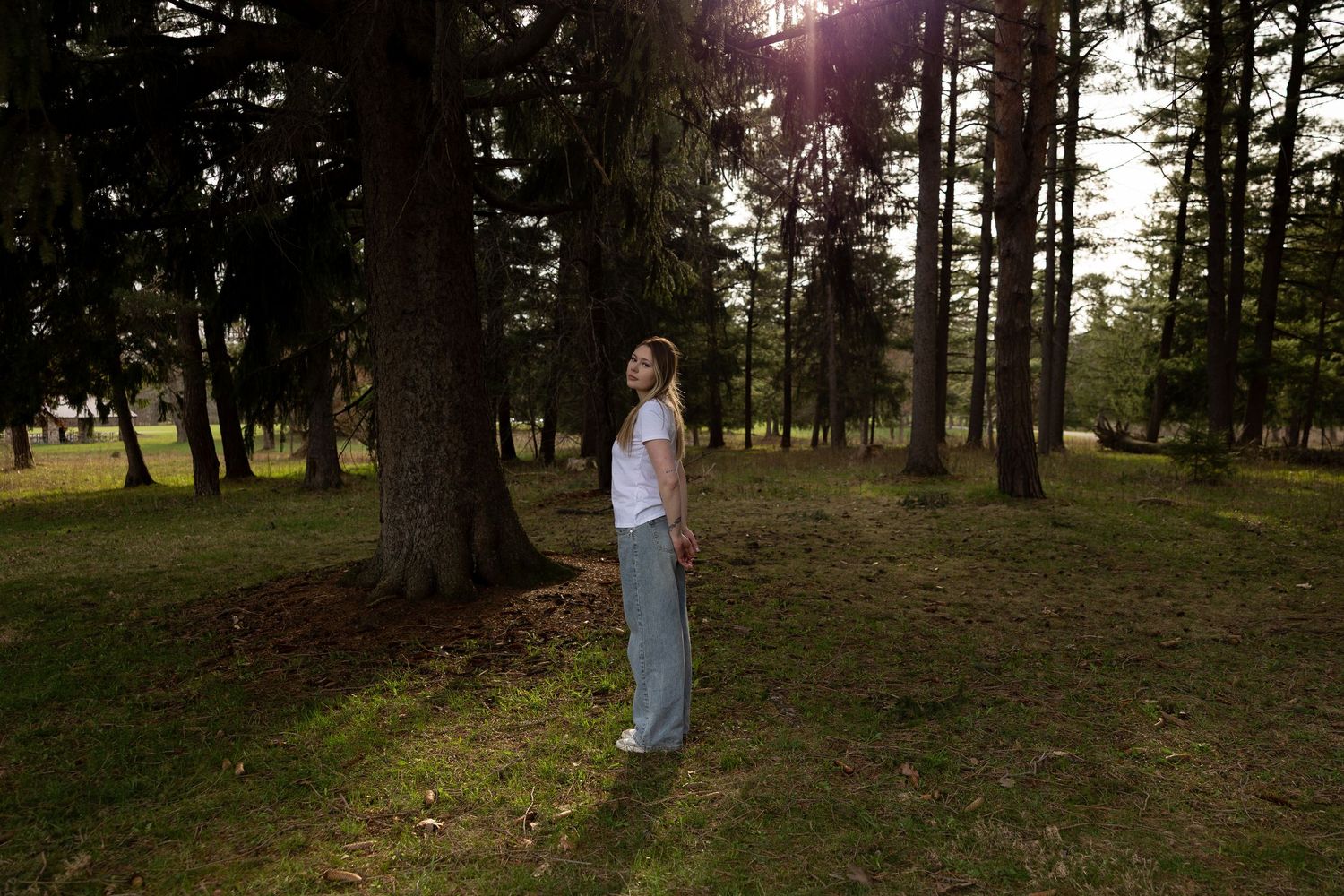 Woman standing among tall trees in a sunlit forest, wearing a white top and light gray pants.