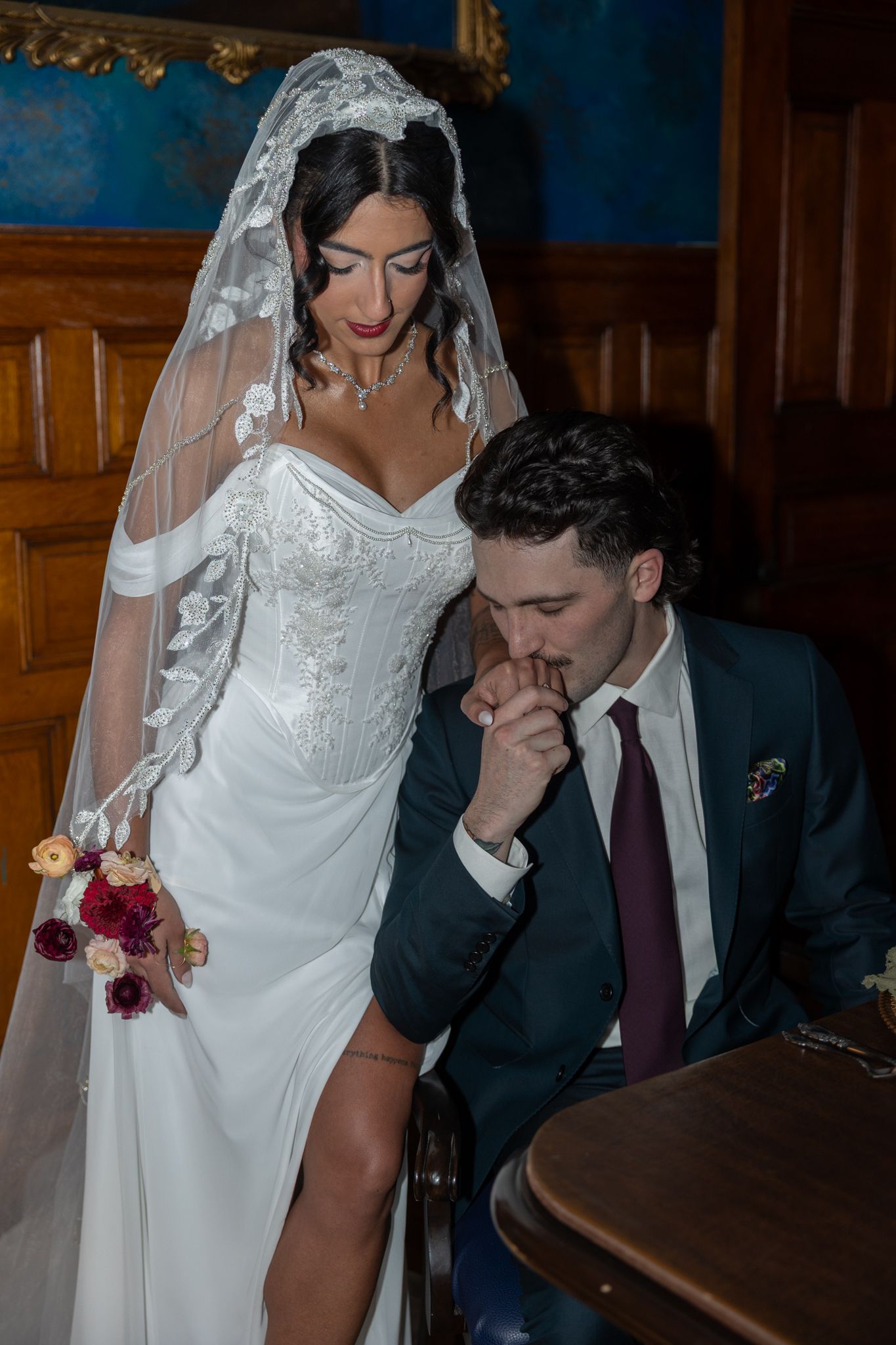 Groom in navy suit kisses bride's hand at wedding ceremony, bride wearing lace veil and white gown holding bouquet.