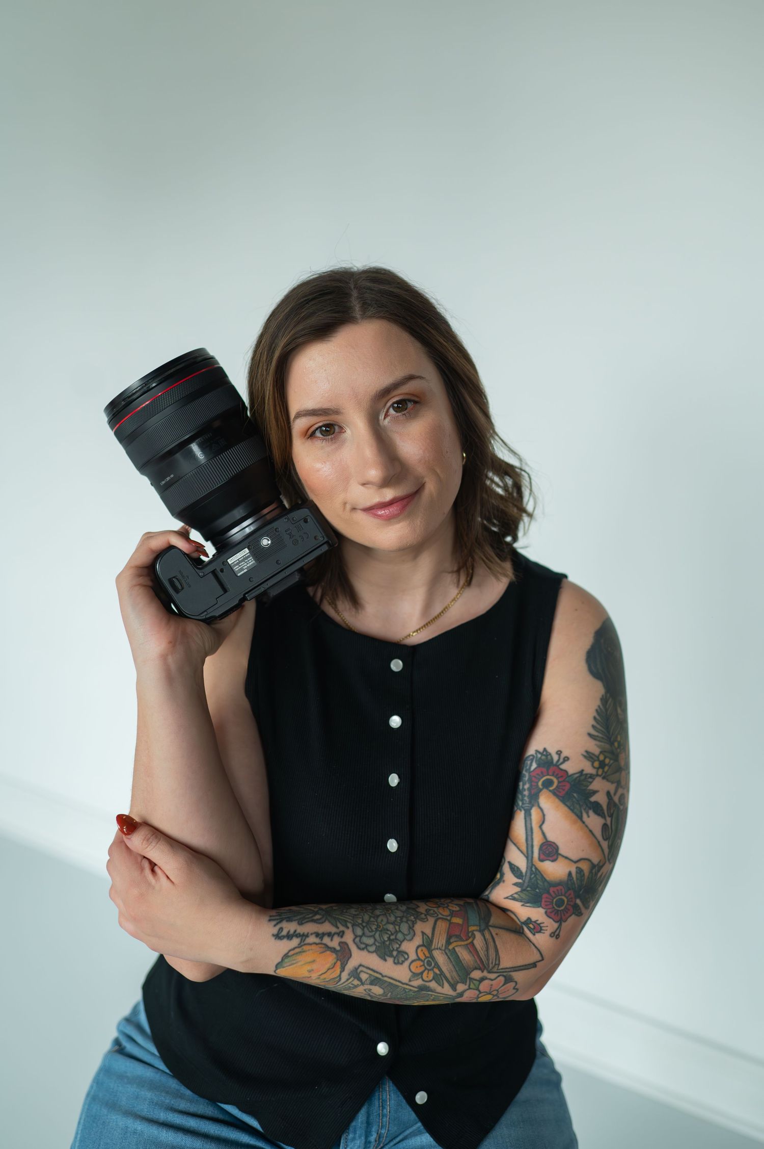 A tattooed female photographer poses confidently holding a Canon camera against her shoulder in a bright studio.