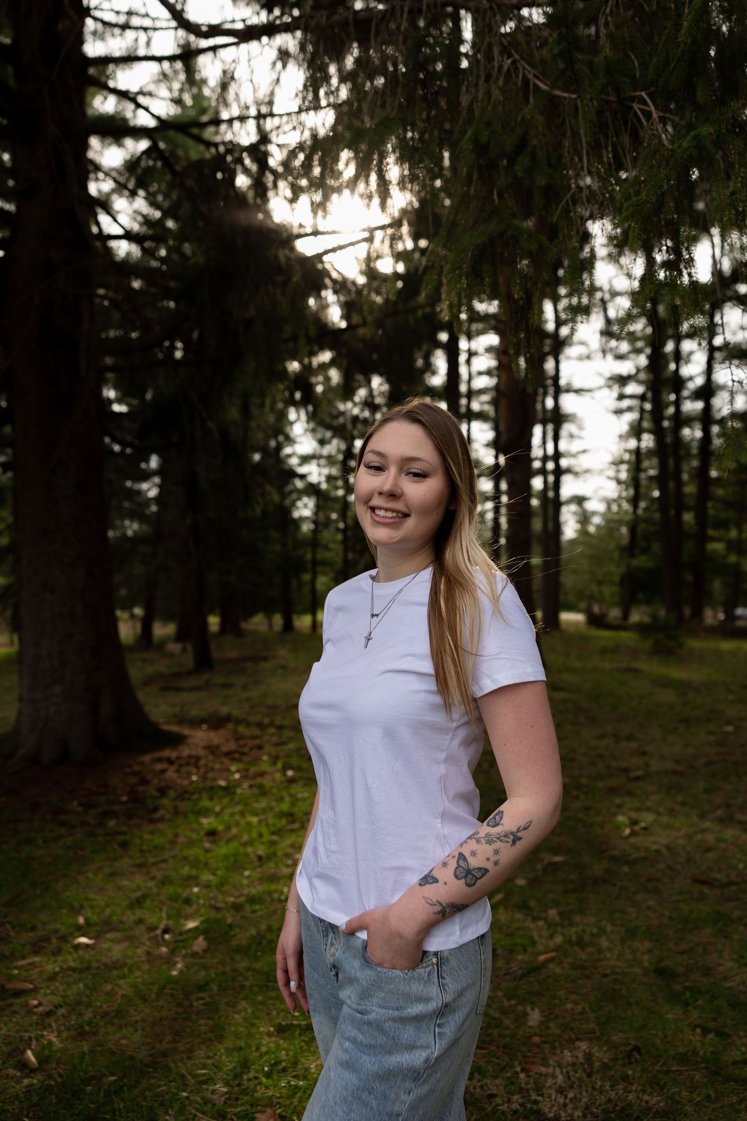 Young woman with tattoos in white t-shirt and jeans smiling in a sunlit forest setting.
