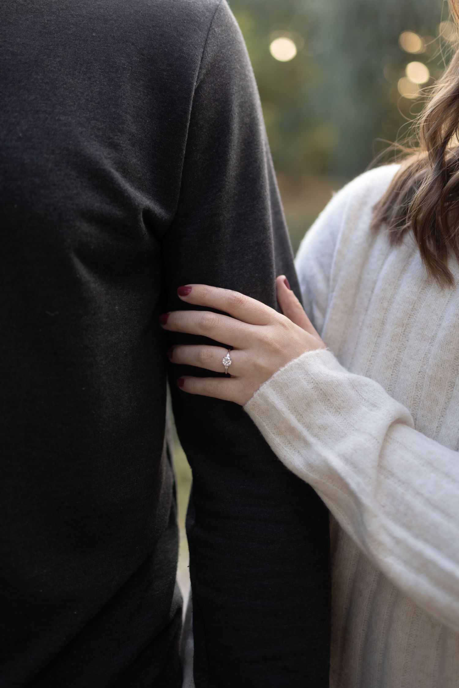 Woman in white sweater wearing diamond engagement ring rests hand on mans dark gray sweater sleeve outdoors.