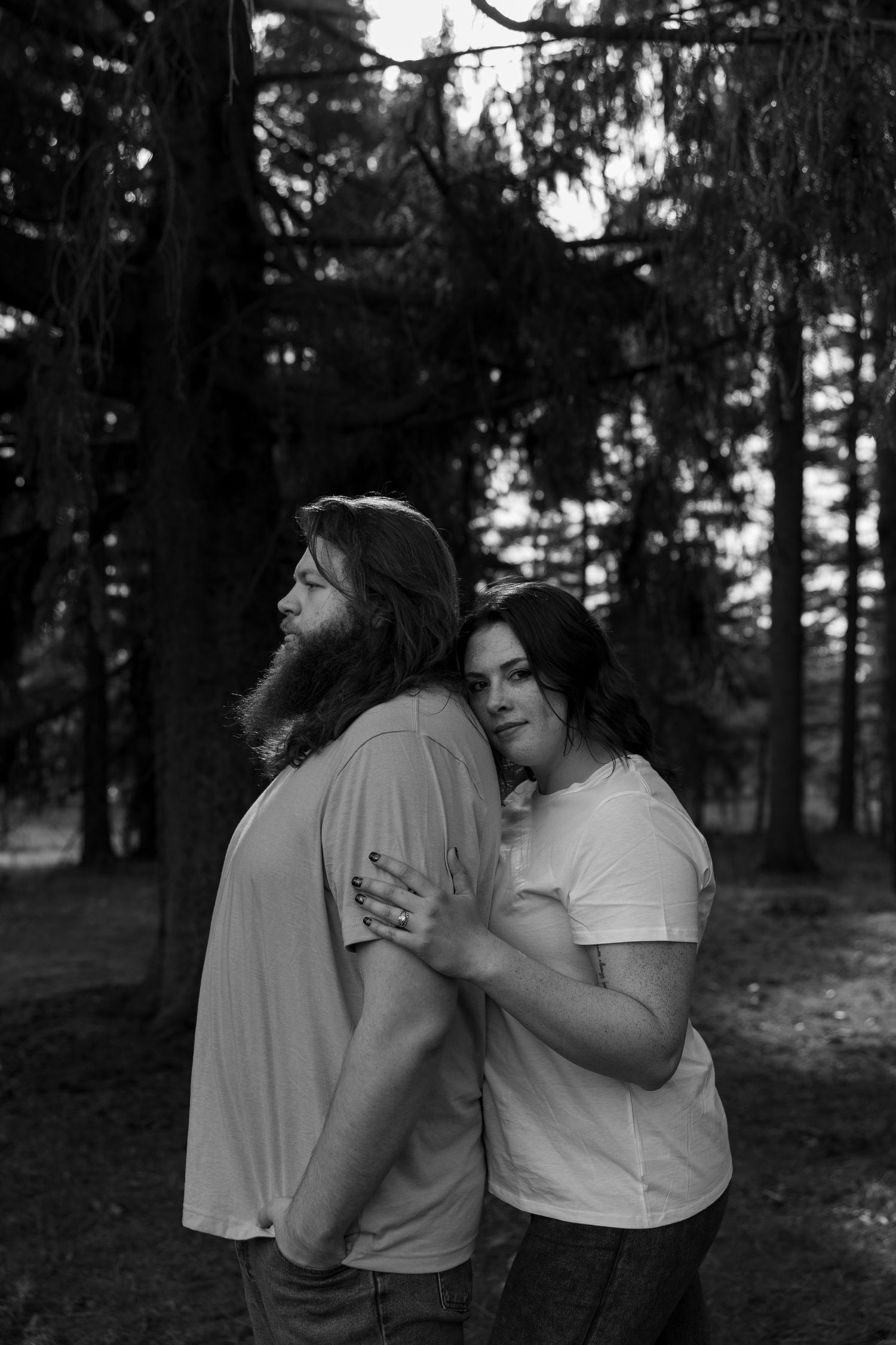 Black and white photo of a couple embracing in a forest, woman hugging man from behind among tall pine trees.