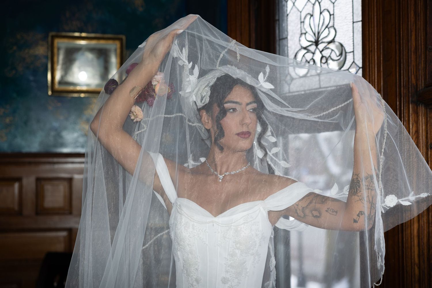 Bride in white wedding dress and veil with tattoos poses elegantly in a vintage-styled room.