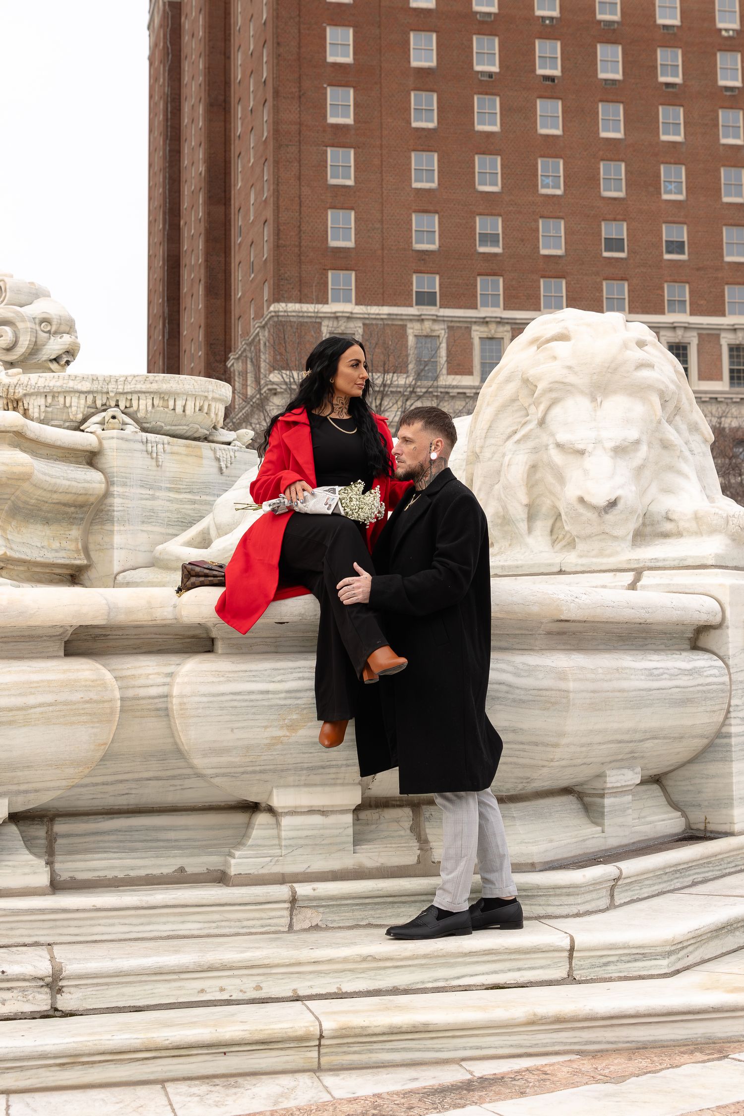 Couple posing at a marble lion fountain in Buffalo, woman in red coat sitting on edge, man in black coat beside her.