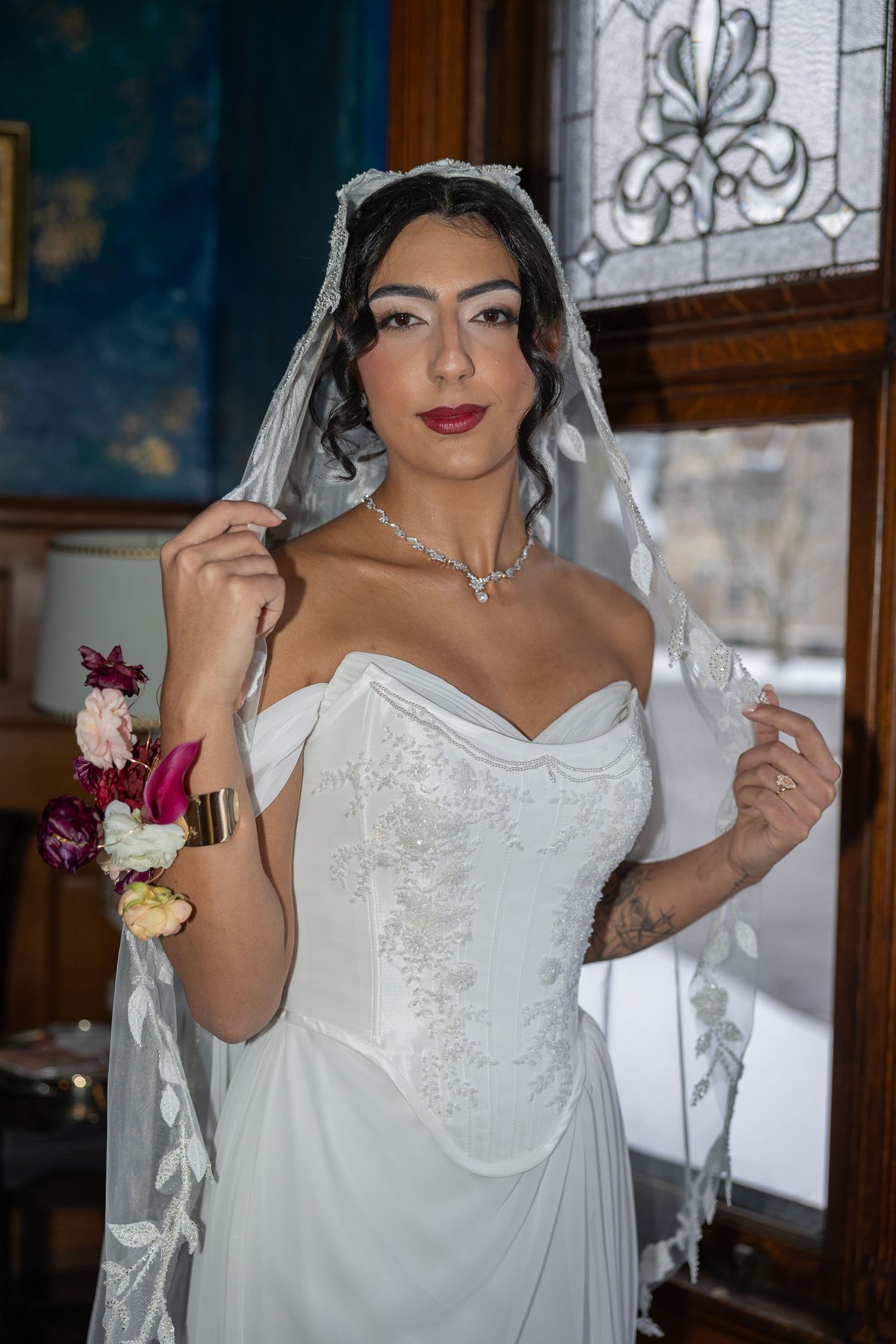 Bride in white off-shoulder gown with lace veil, floral wrist corsage, and diamond necklace posing near stained glass window.