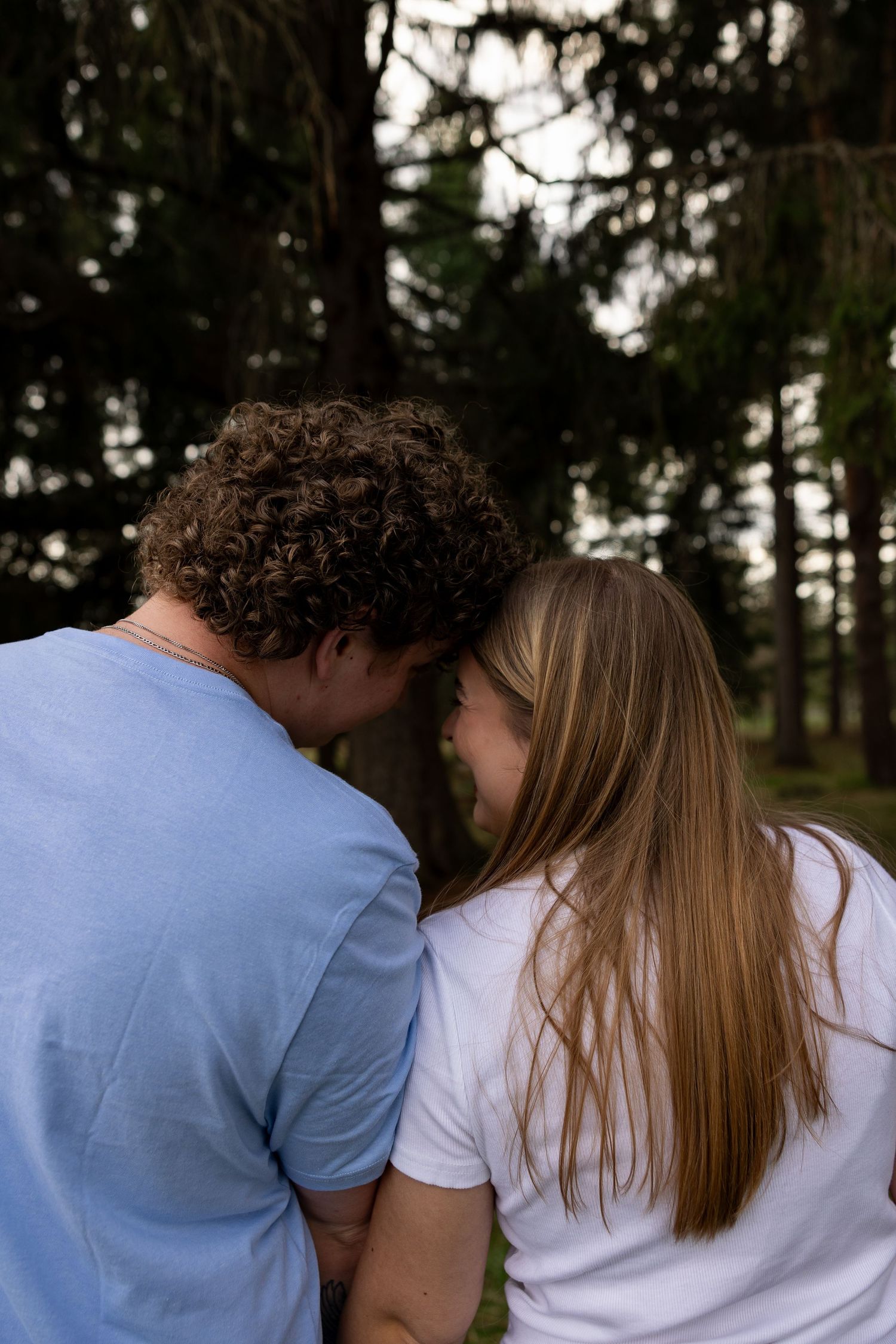A couple sharing an intimate moment outdoors, leaning heads together surrounded by tall green trees in a forest.