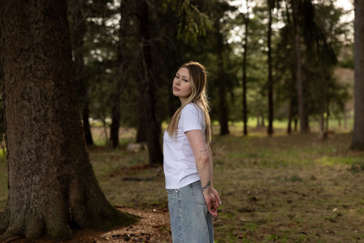 Young woman in white t-shirt and jeans standing among tall trees in a forest, looking over her shoulder.