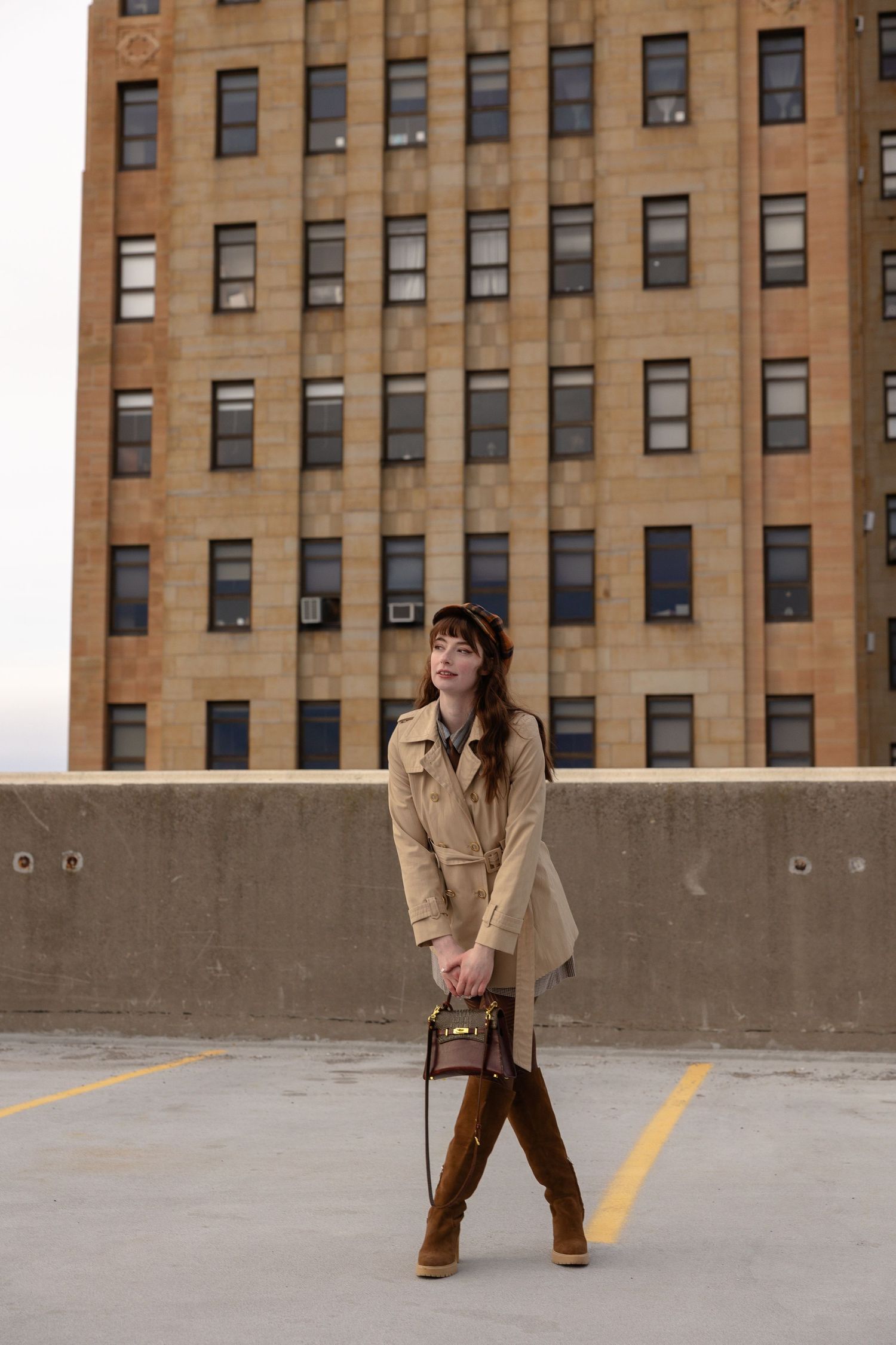 Woman in beige trench coat and brown boots poses on rooftop parking lot with urban building backdrop.